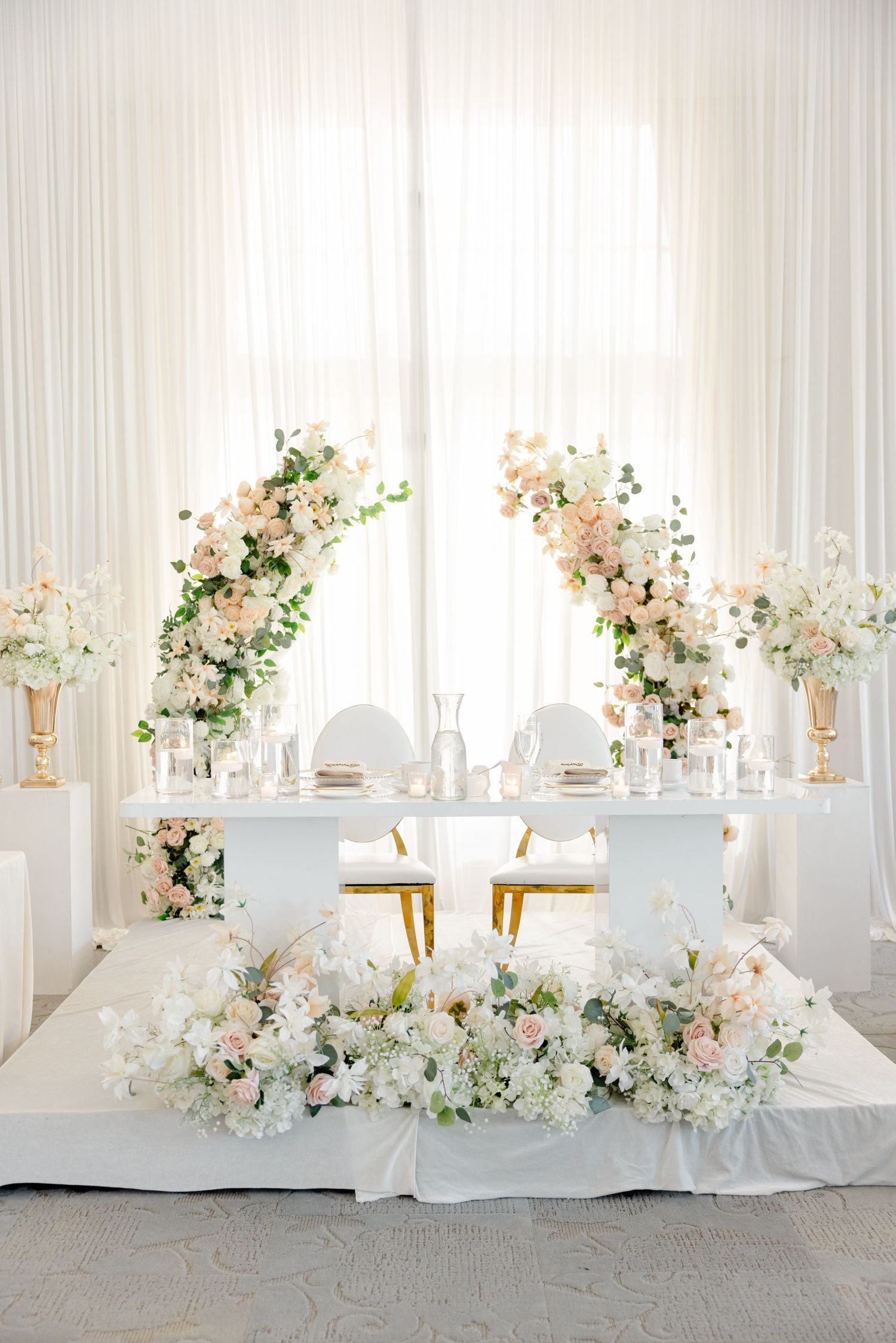 Elegant white wedding reception table setup with floral arrangements and gold accents in a bright modern venue.
