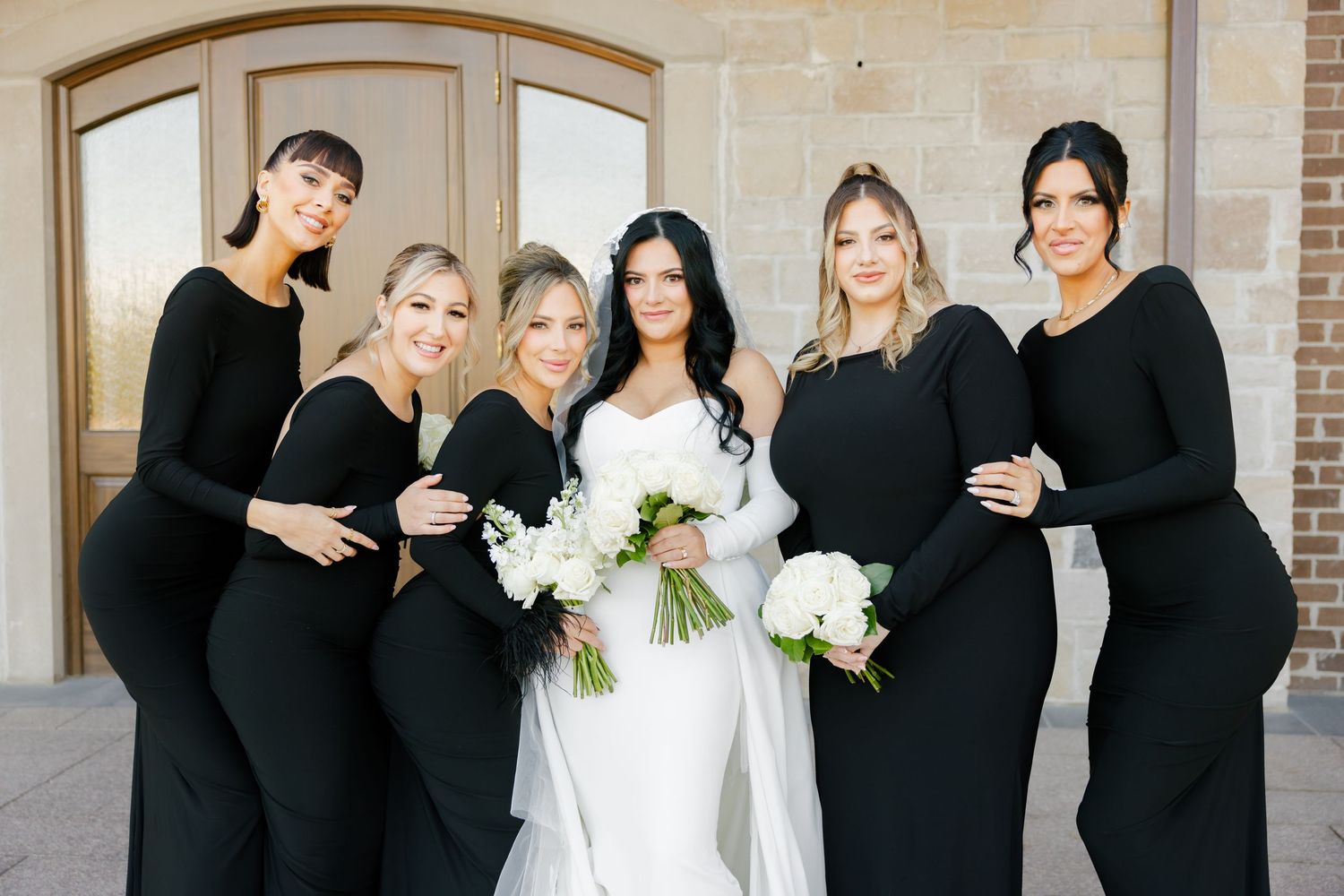 Bridal party in sleek black dresses poses with white floral bouquets.