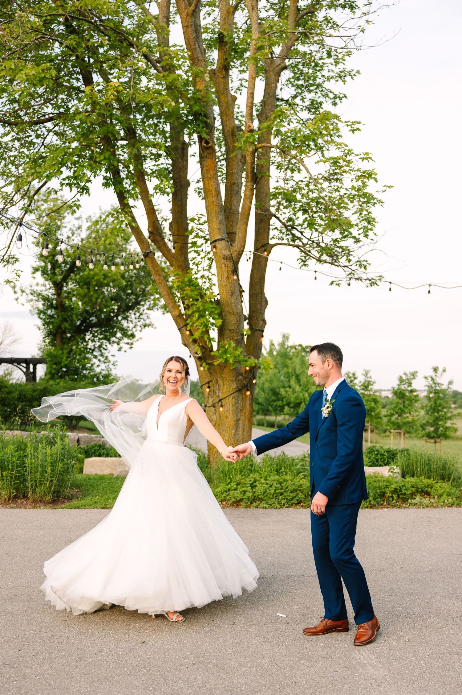 A bride's veil flows in the breeze as she dances with her partner under a majestic tree.