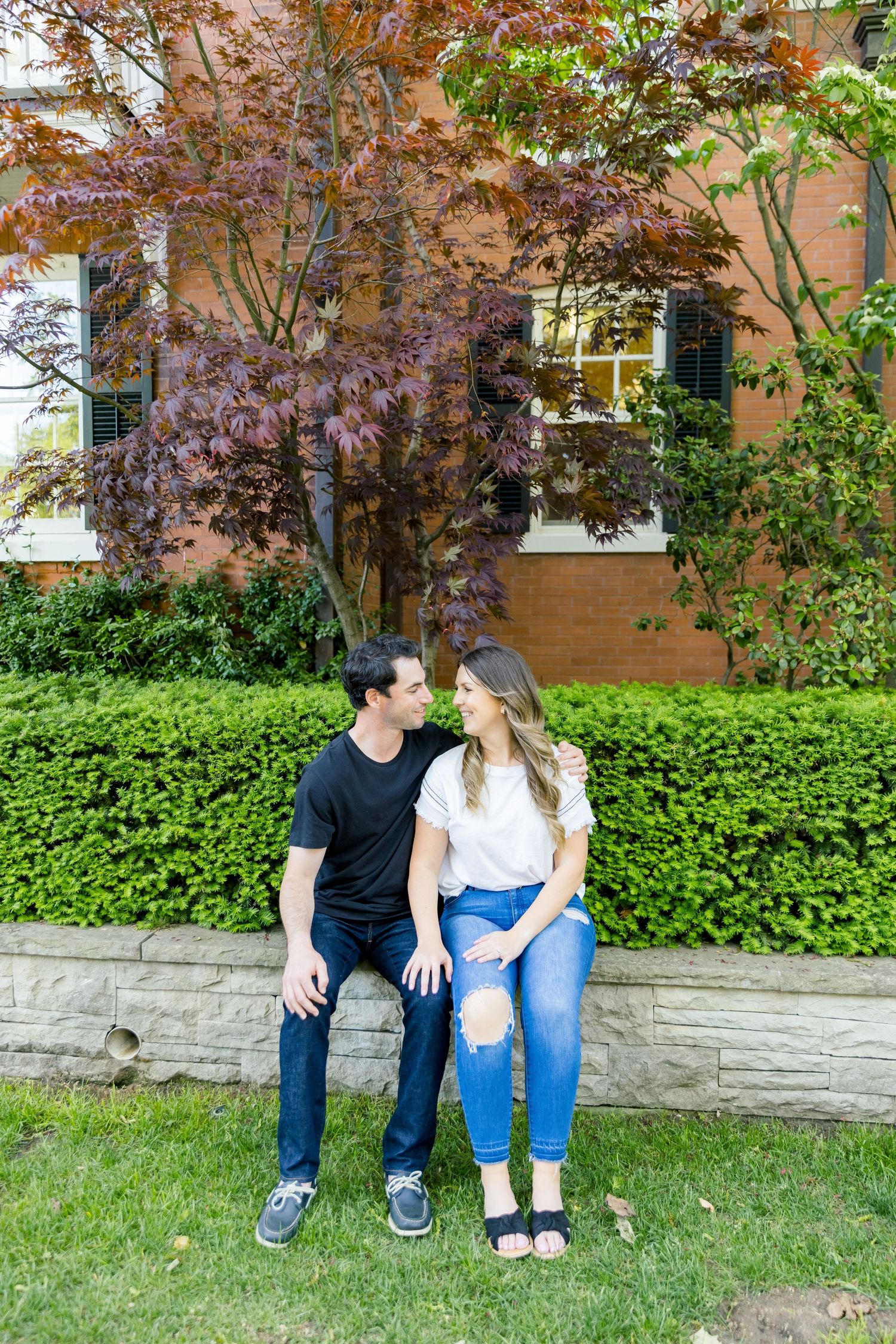 A couple sits on a stone wall beneath a flowering tree with red leaves and a brick building in the background.