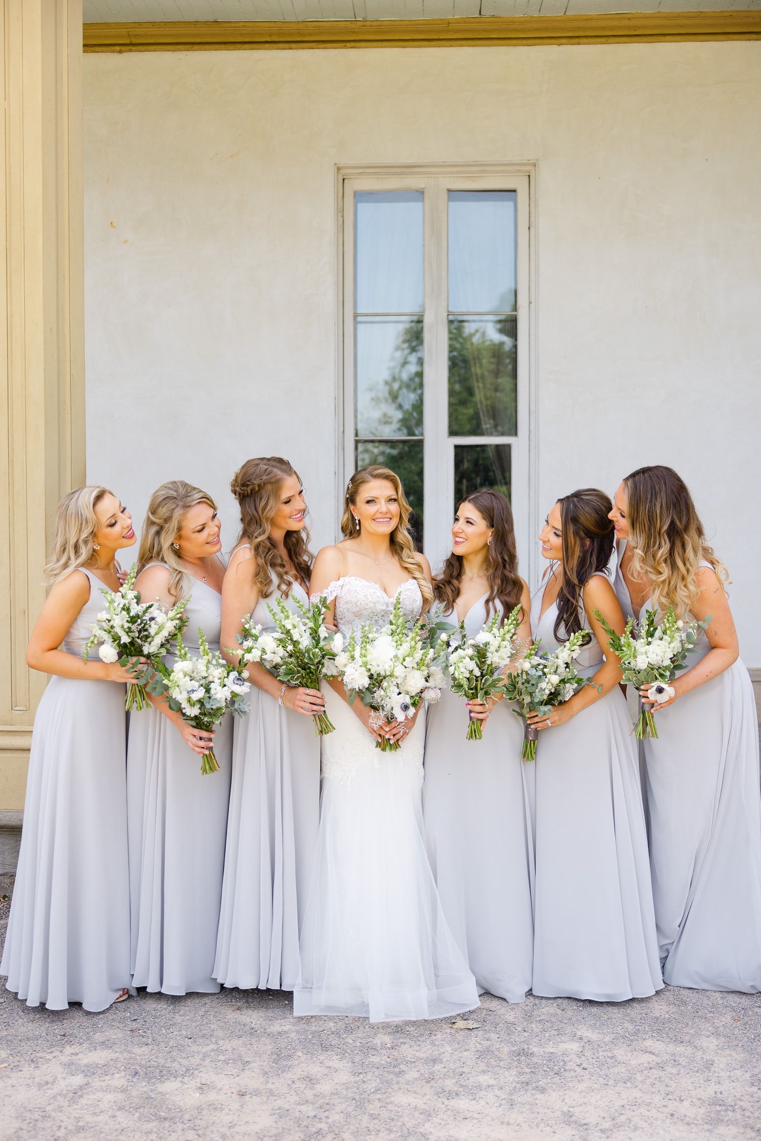 Bridal party in elegant gray gowns pose with white floral arrangements against white building.