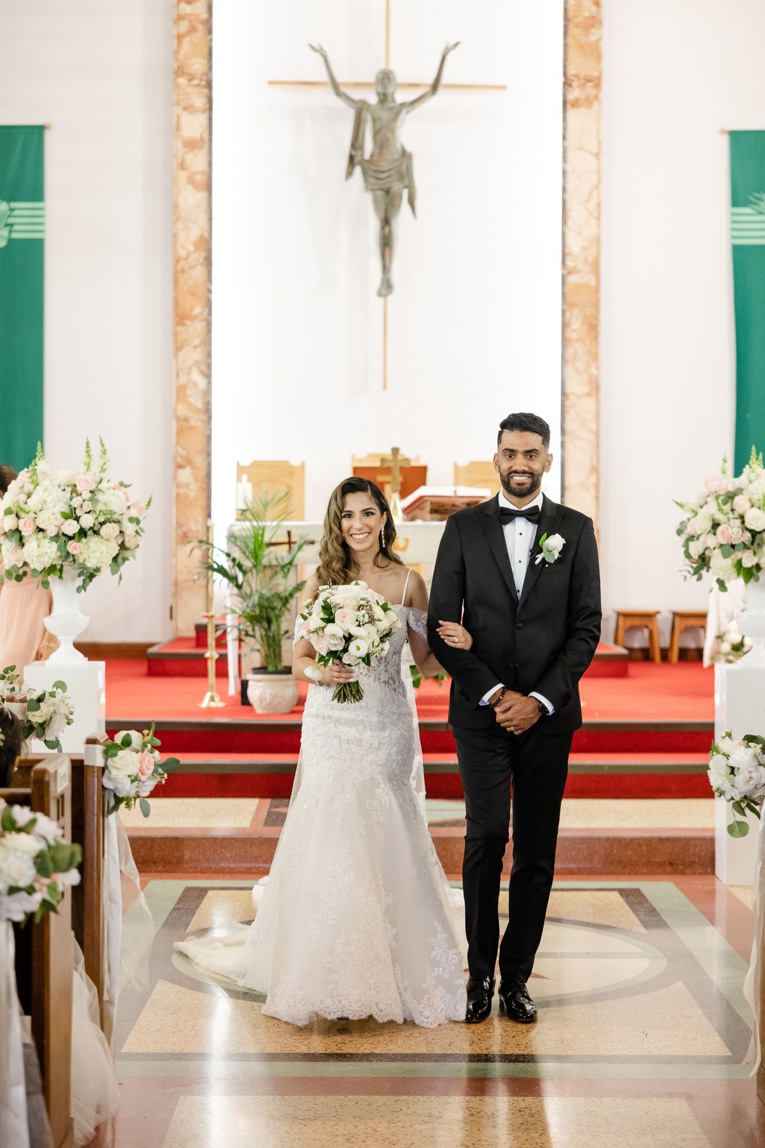 A formal wedding portrait in a Catholic church with white floral arrangements and green banners.