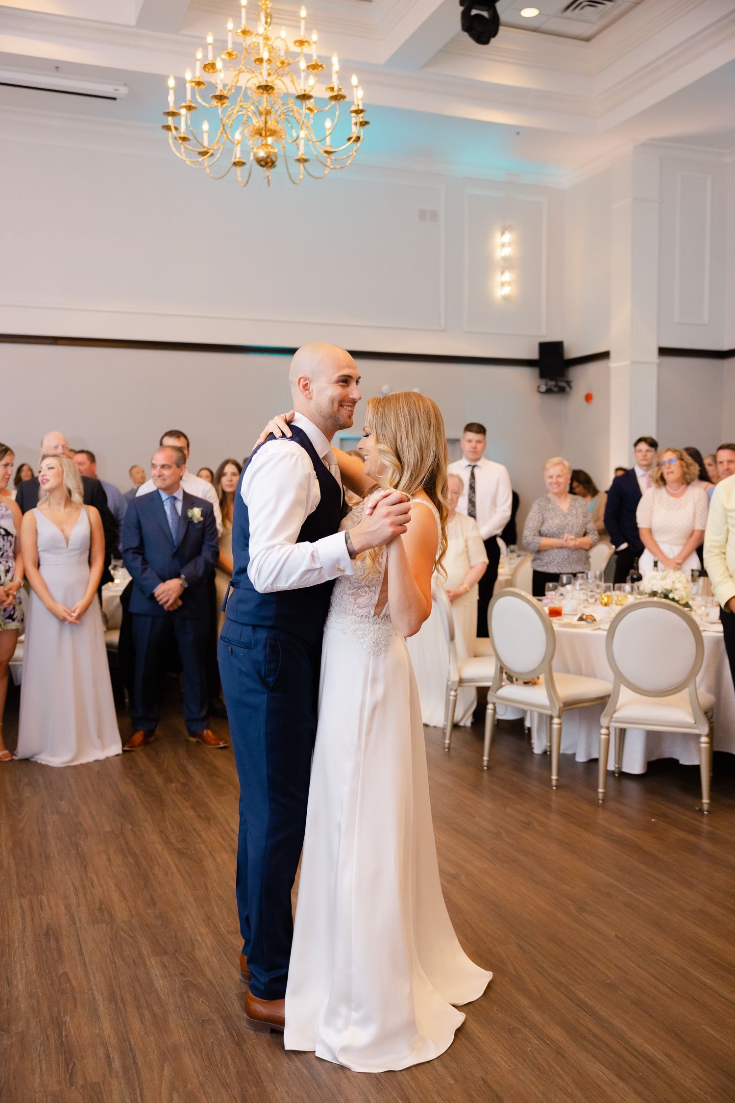 Couple shares their first dance at wedding reception under elegant chandelier as guests look on.
