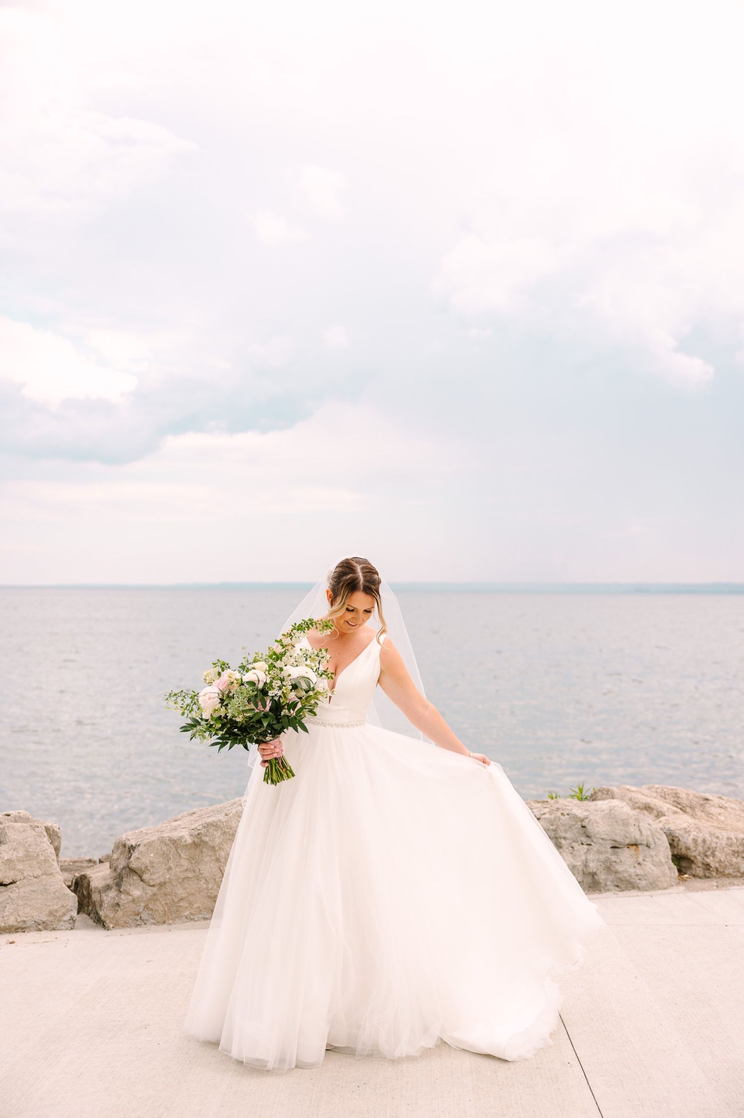 A bride in a flowing white dress holds a bouquet while posing by the ocean on a cloudy day.