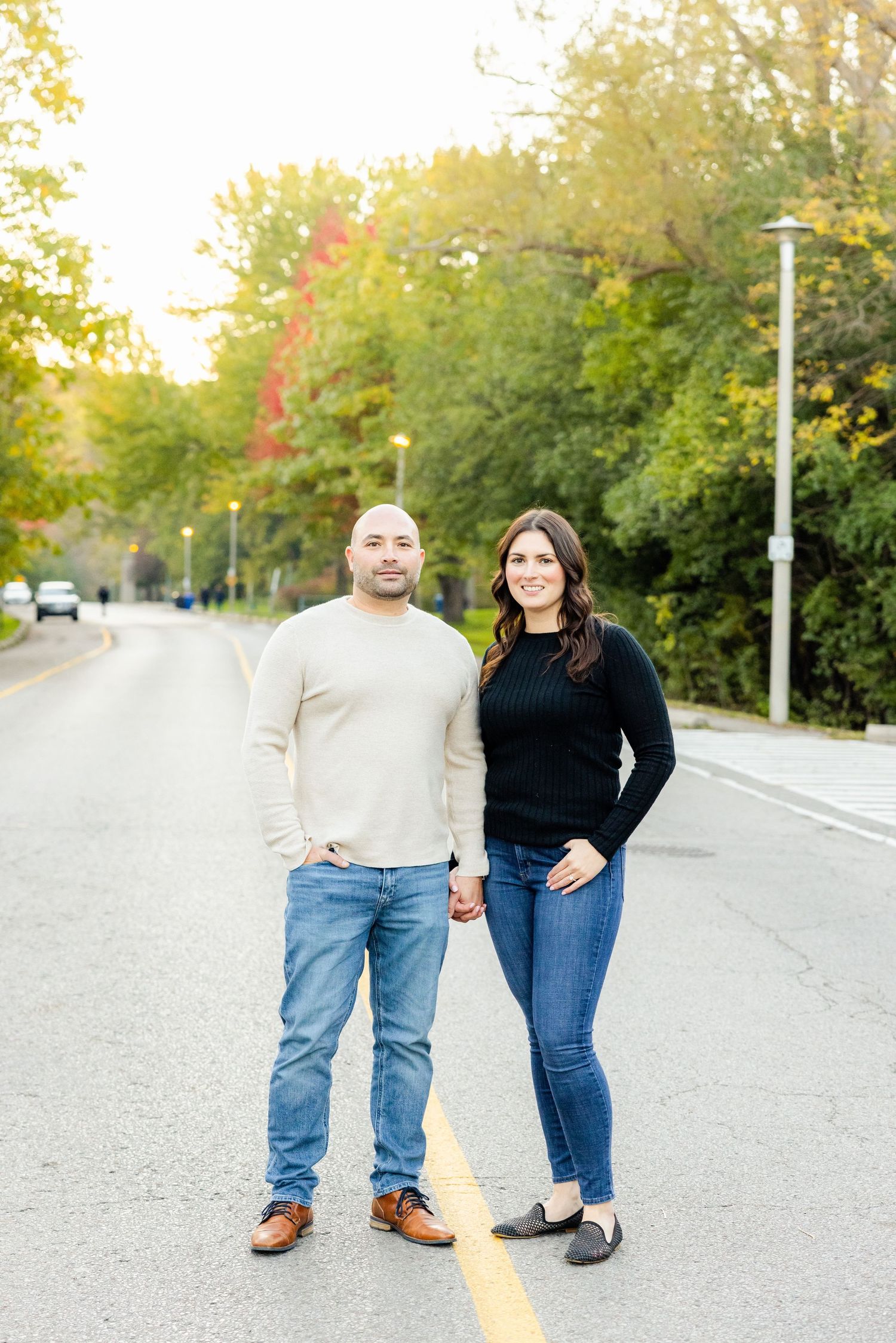 A couple poses together on a road surrounded by fall foliage wearing casual sweaters and jeans.