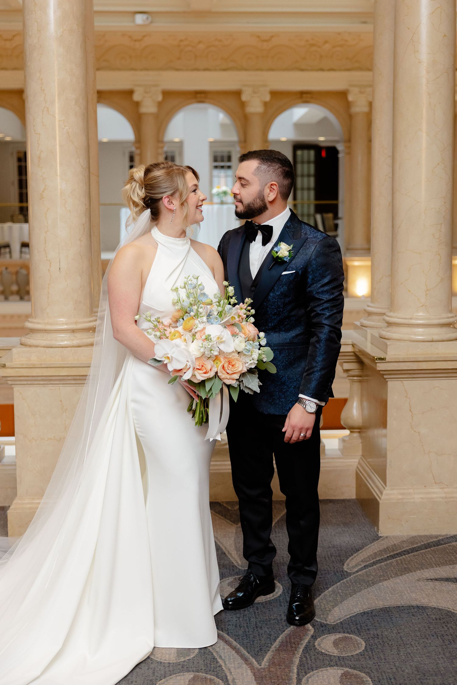 A romantic wedding first look moment between a bride in white and groom in black suit within an elegant columned hallway.
