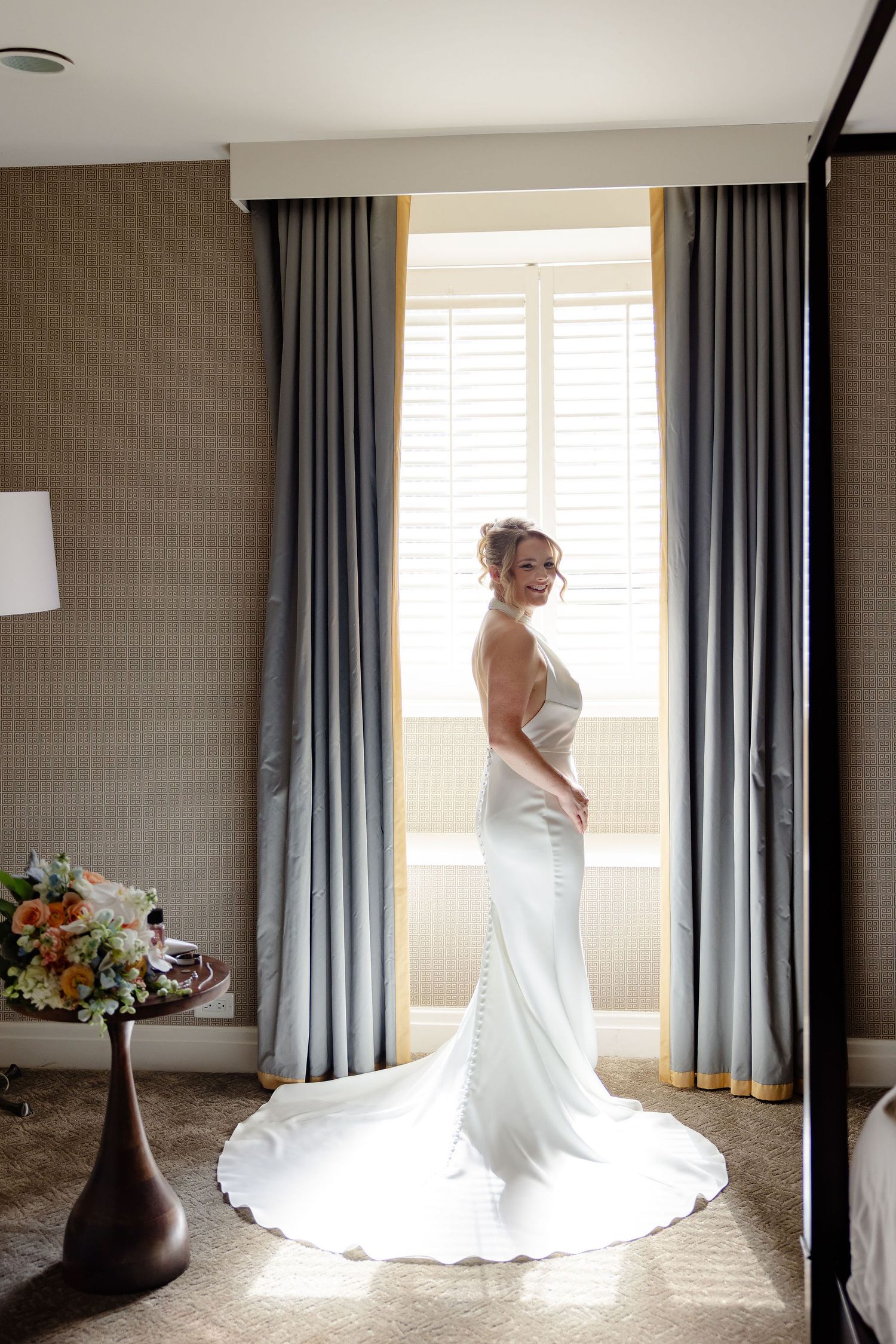 Sunlight streams through windows illuminating a flowing white wedding dress against grey curtains.