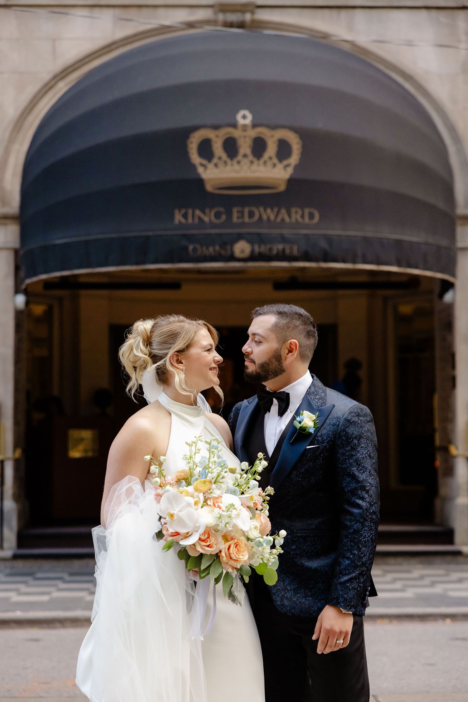 A couple shares a romantic moment holding bouquets of peach and white flowers outside a hotel entrance.