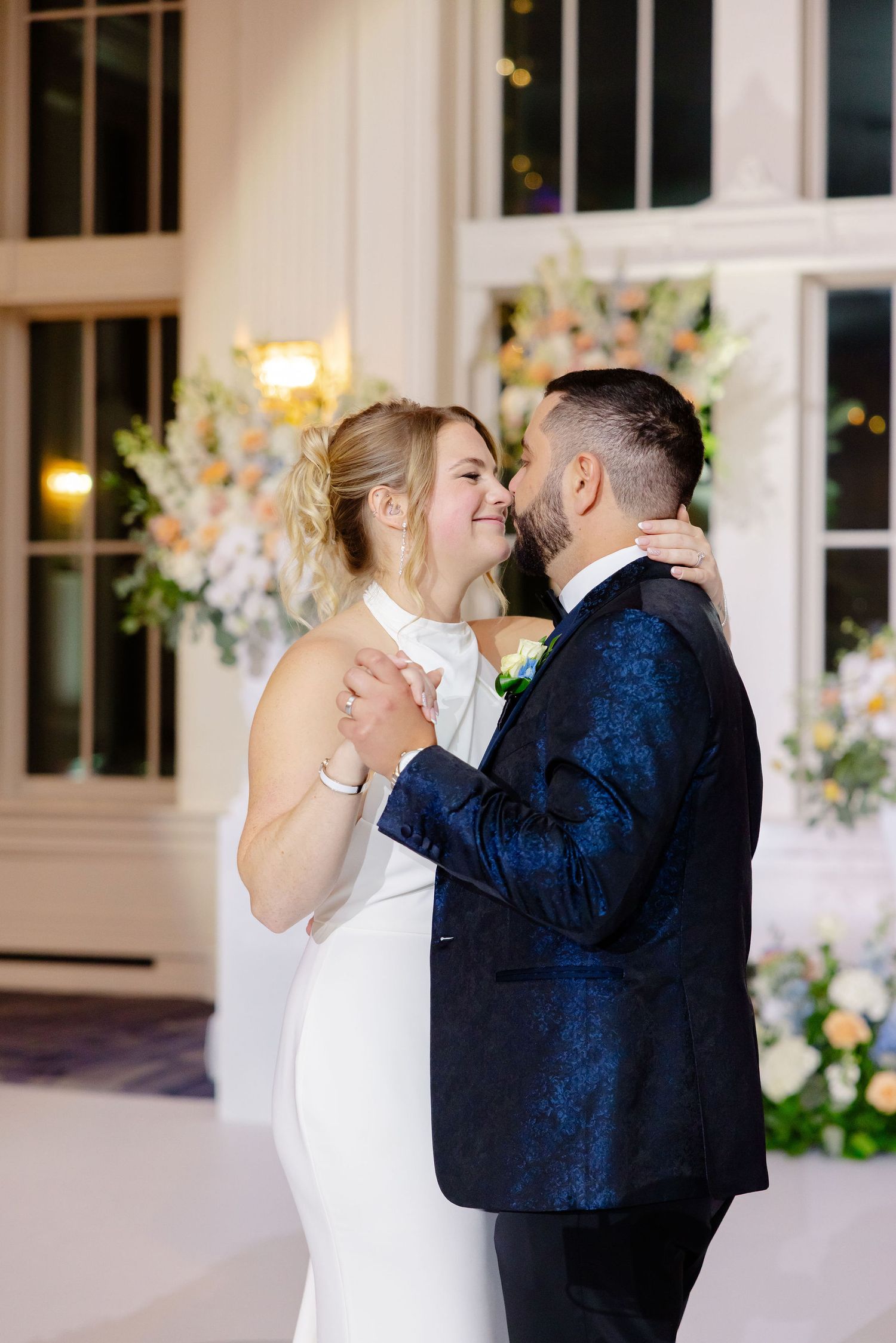Intimate wedding moment with bride in white dress and groom in navy tuxedo against floral backdrop.