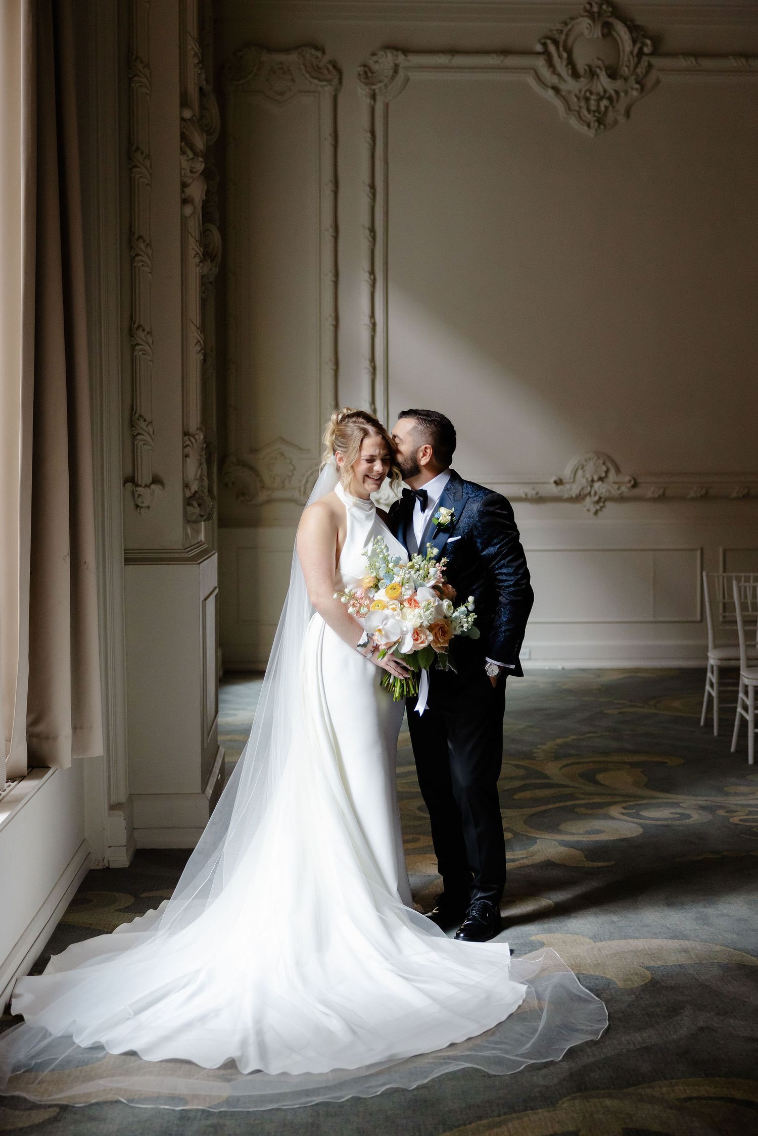 Elegant wedding couple embraces in ornate room with decorative wall moldings and long flowing veil.