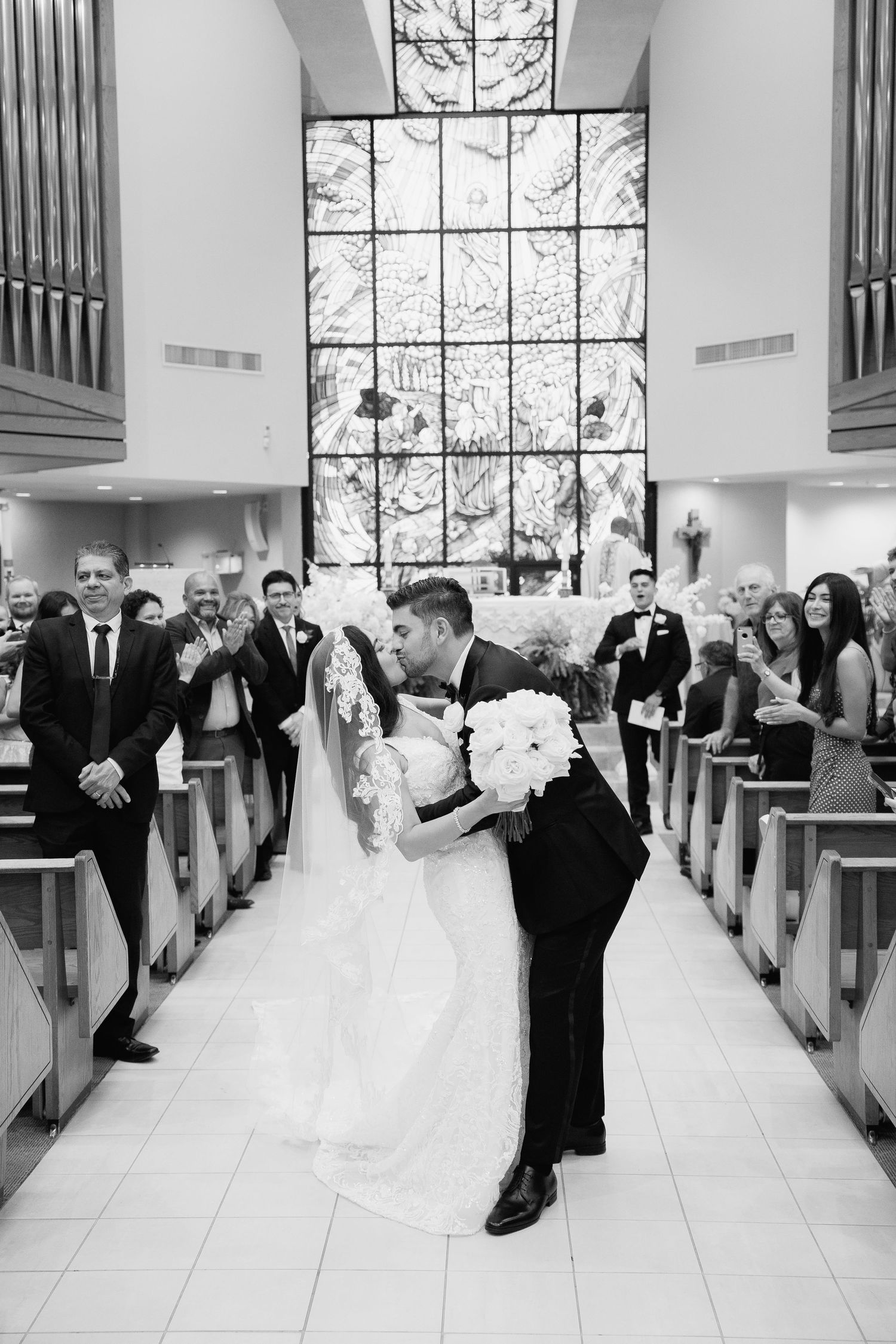 Romantic wedding kiss in church aisle with guests watching from pews.