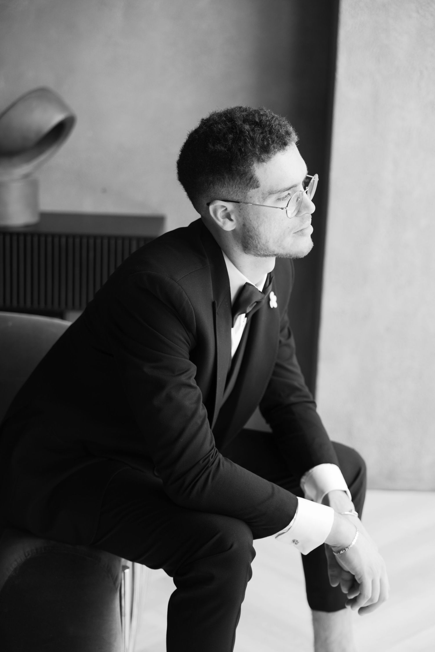 A black and white portrait of a contemplative person in a suit and glasses sitting in profile.