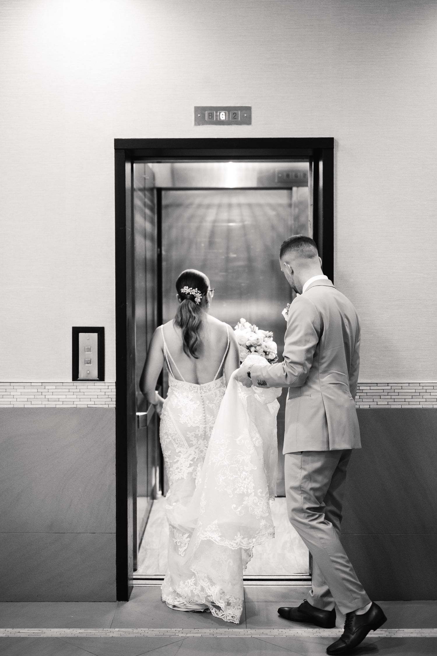 Black and white photo of couple entering elevator with long wedding dress train.