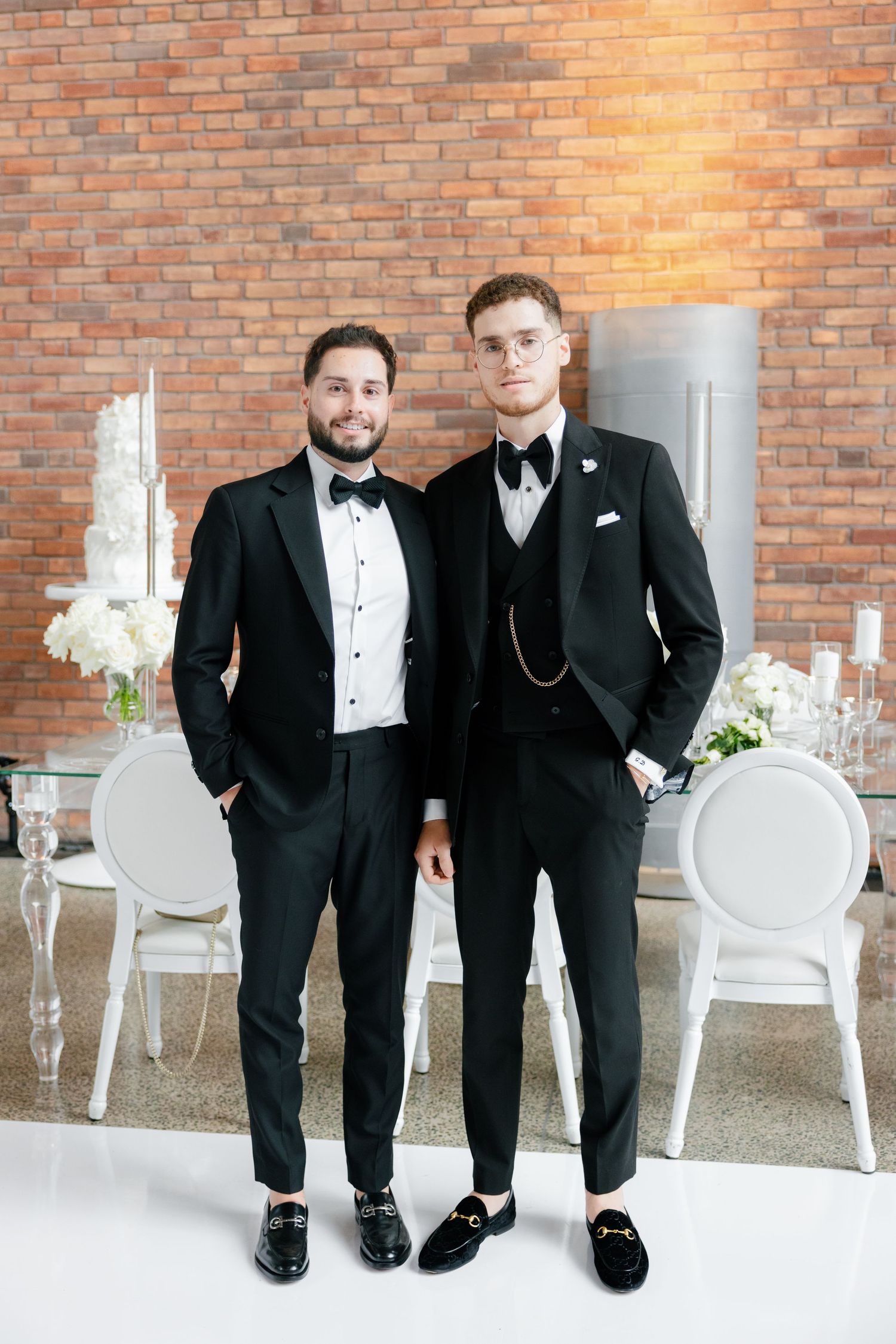 Two people in classic black tuxedos pose together at formal event with brick wall background.