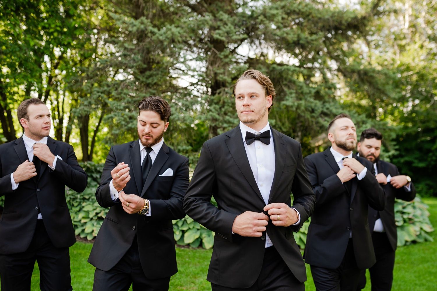 Groomsmen in black tuxedos and bowties adjust their attire while standing outdoors in a garden setting.