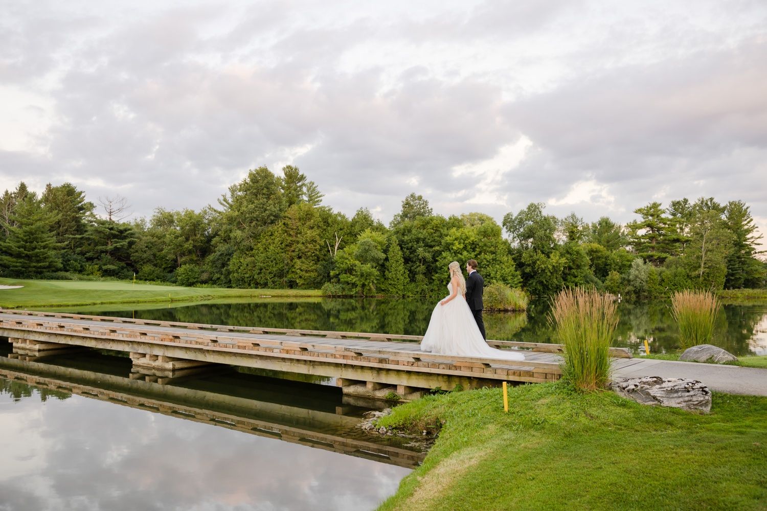 White wedding dress silhouette reflected in calm water by wooden dock surrounded by lush greenery.