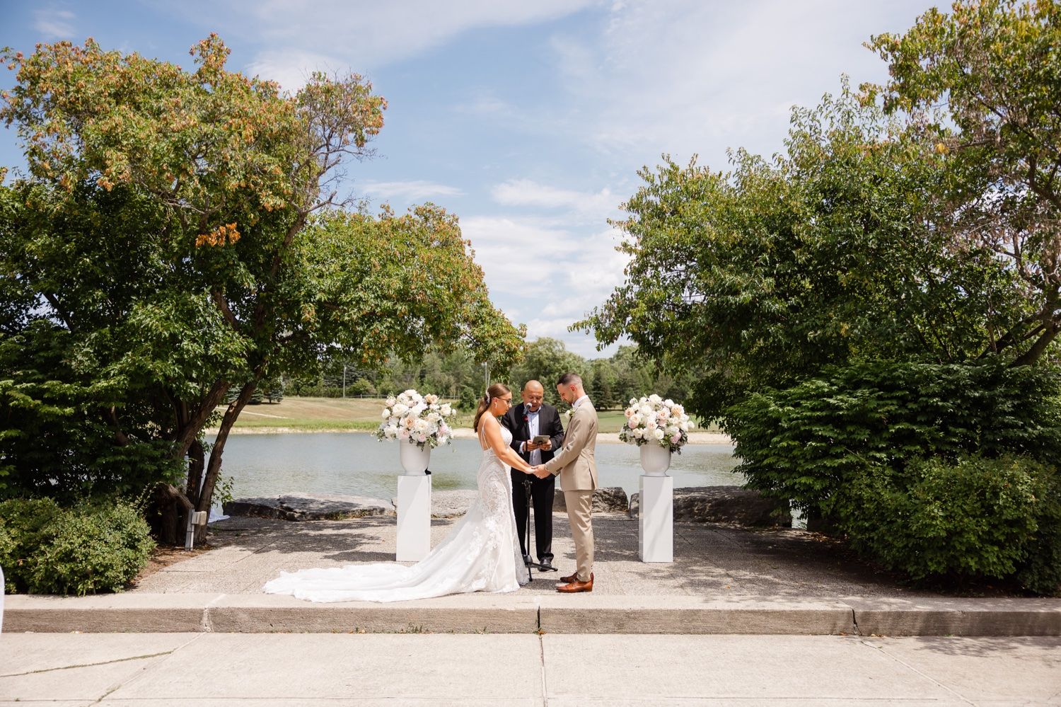 Lakeside wedding ceremony setup with white floral pedestals and natural garden surroundings.