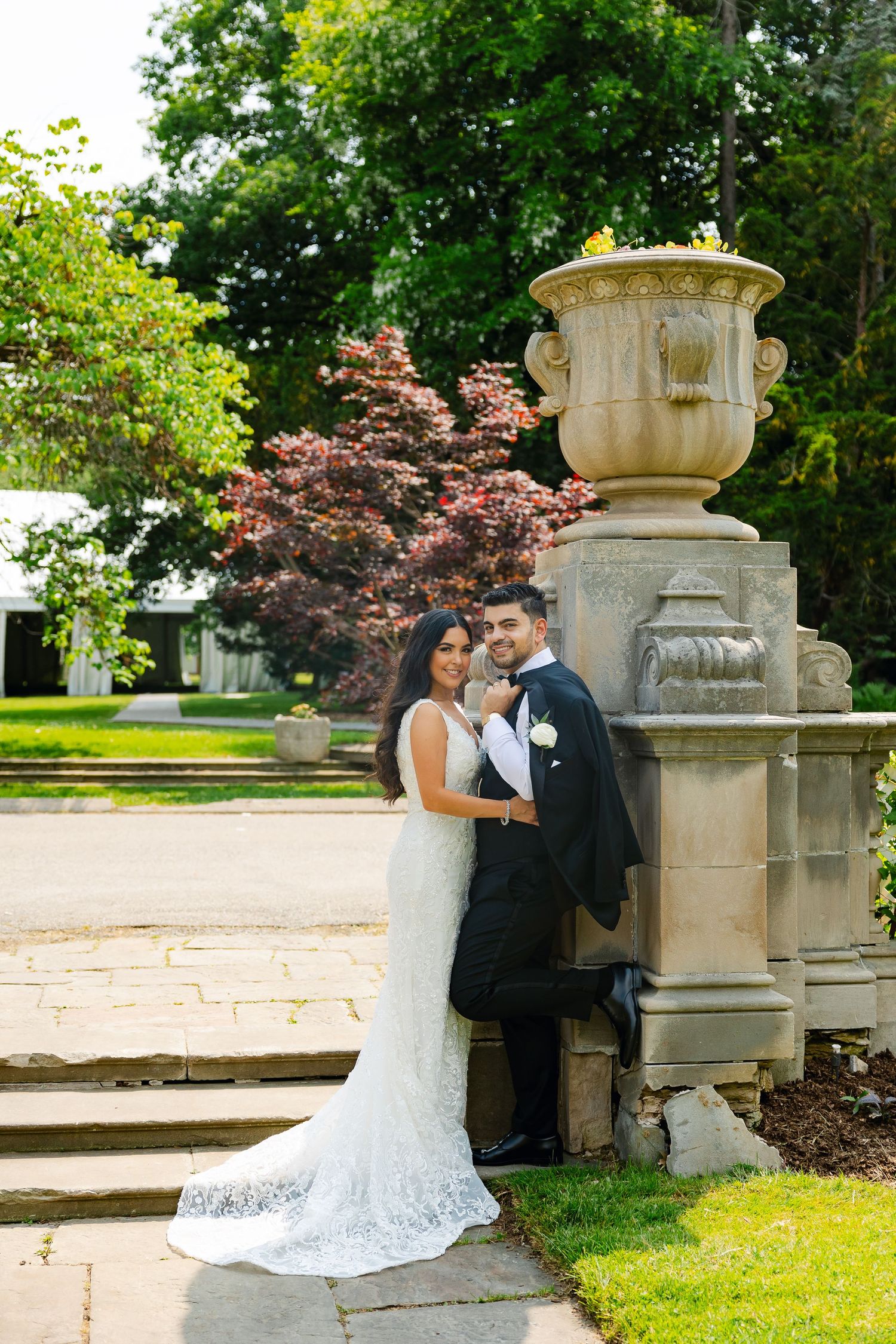 Newly married couple stands by stone urn pedestal with colorful garden foliage in background.