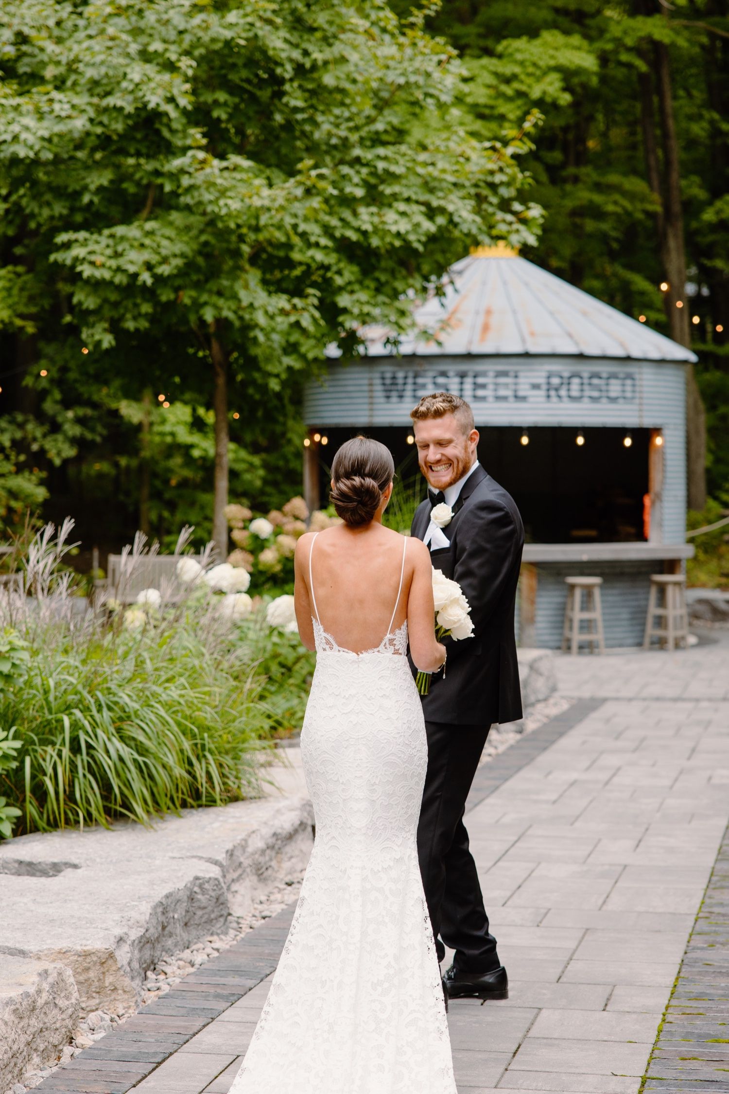 A bride in a fitted white dress and groom in black tuxedo share a moment on a stone path near Westeel-Rosa venue.