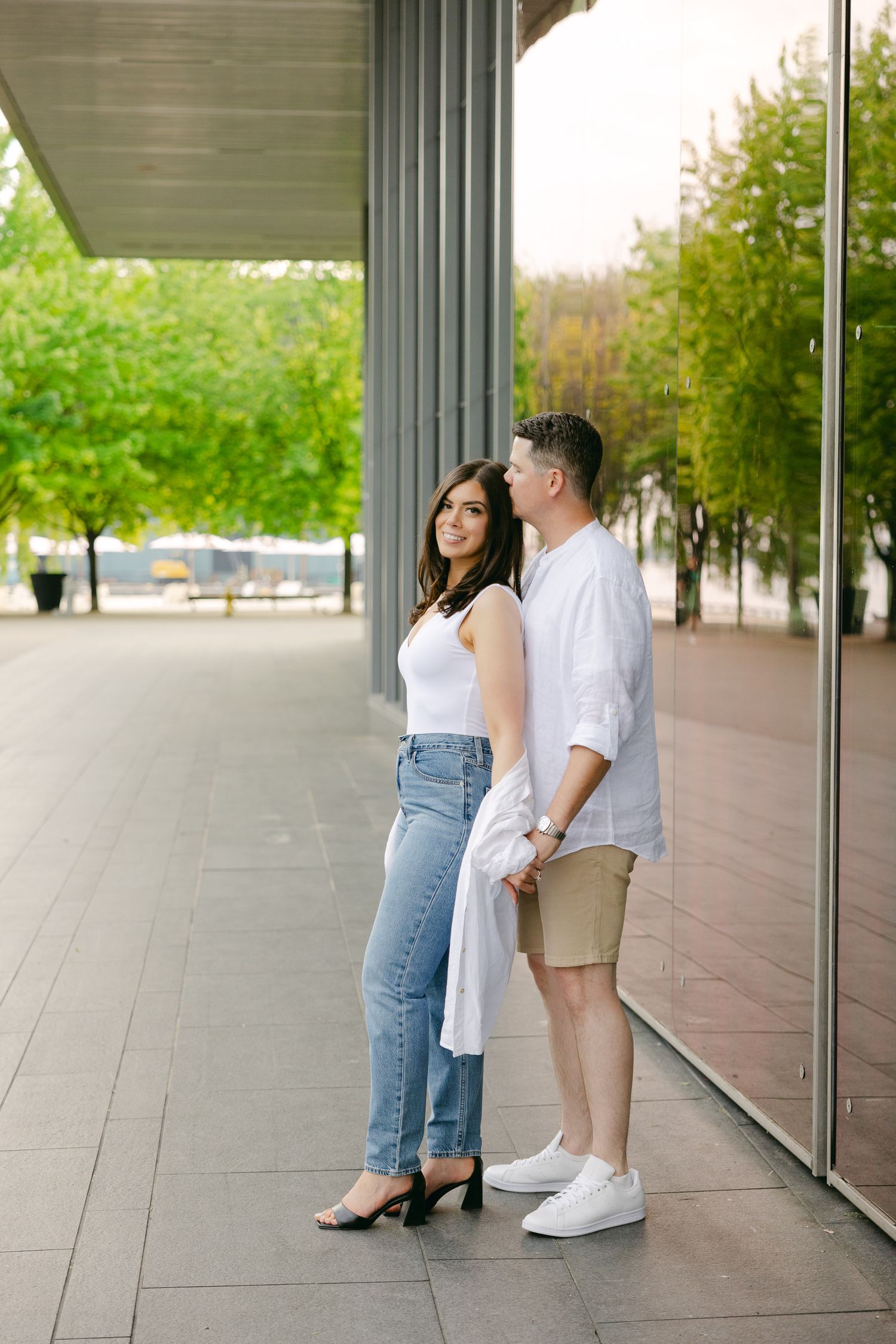 A couple stands together against reflective glass windows in an outdoor covered walkway.