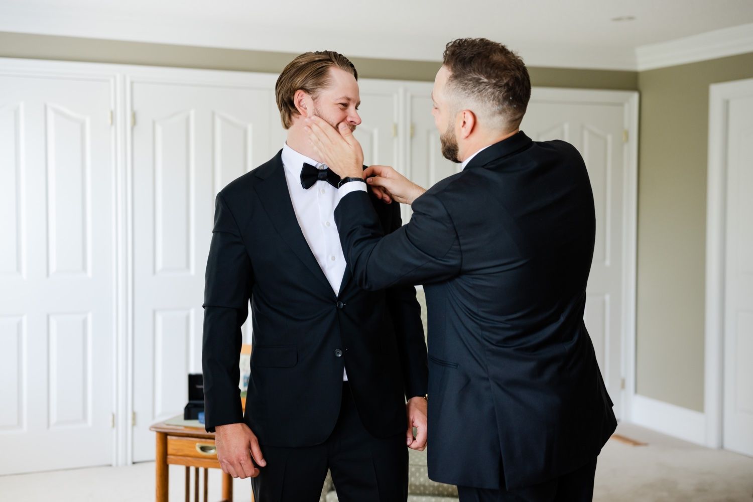 Two people in tuxedos helping each other with bow tie adjustments in a light filled room.