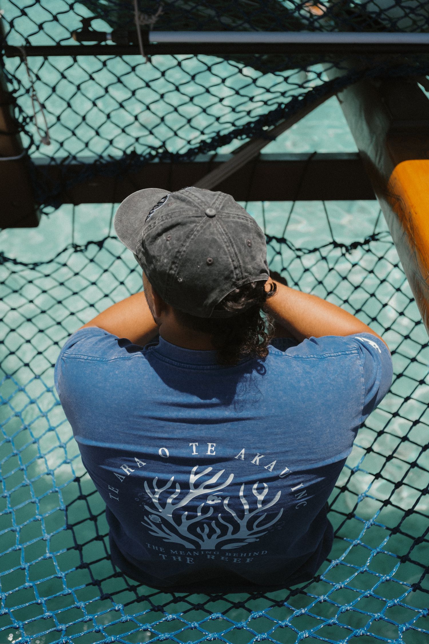 Person in blue graphic tee and gray cap sits on netting, viewed from behind, looking out over water.