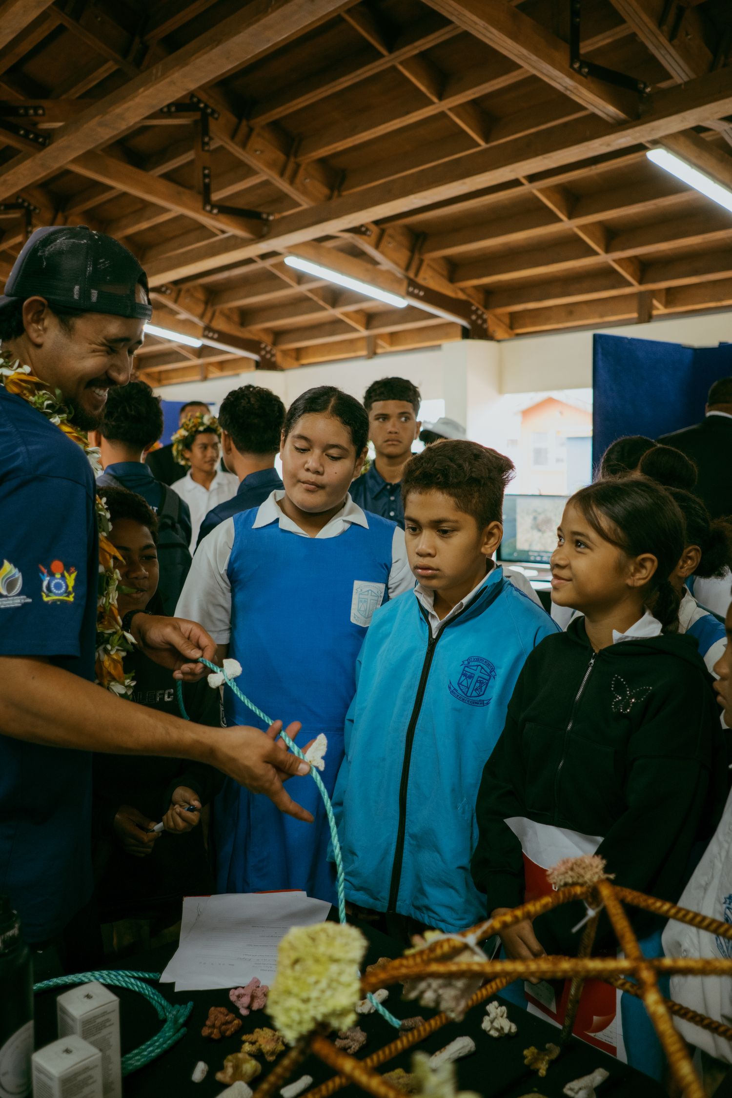 Students gather around a display table at an educational fair, listening to a presenter wearing a floral lei.