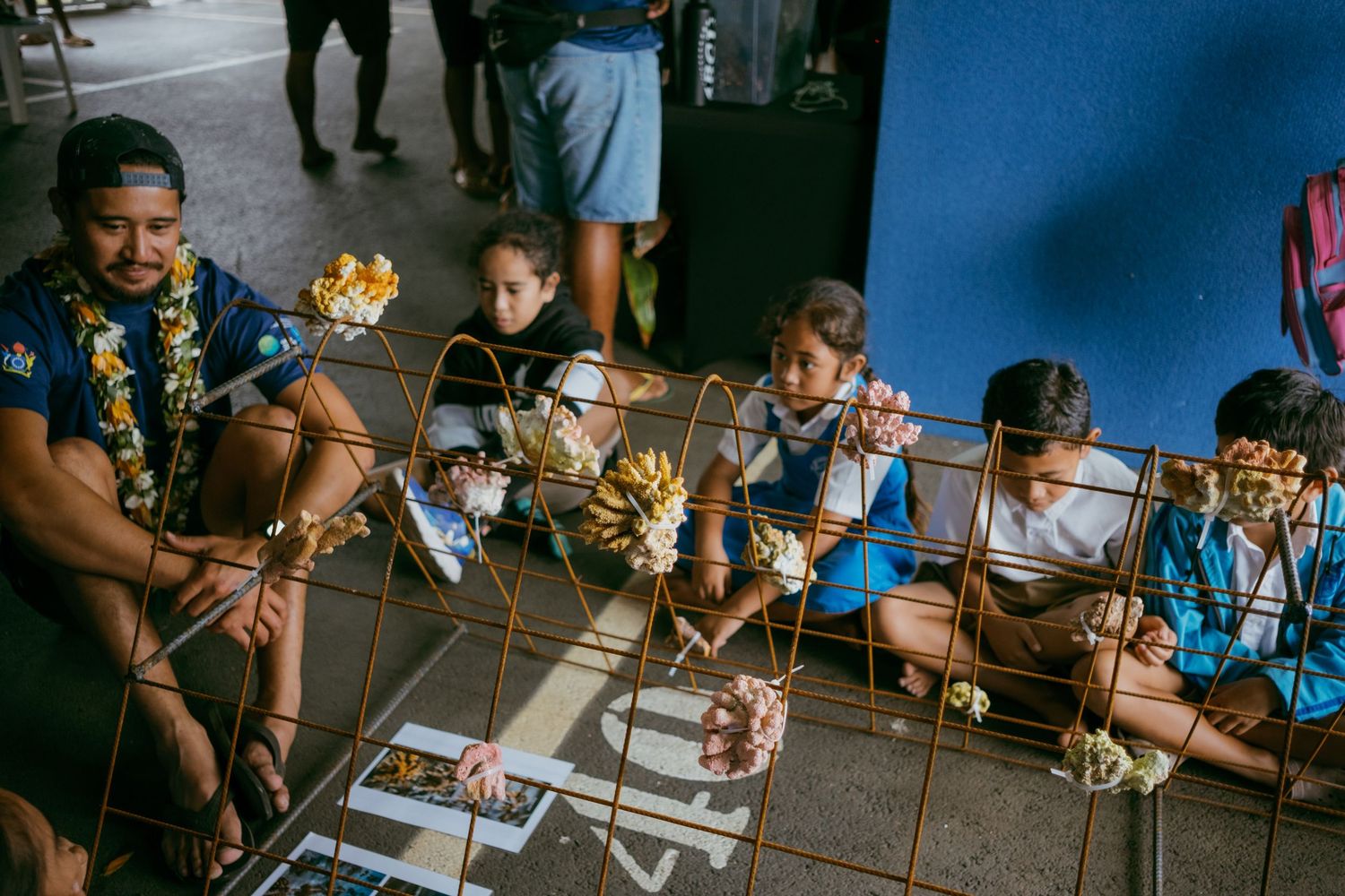 Children in traditional attire sit behind a bamboo fence at a cultural event, accompanied by an adult in a blue floral outfit.