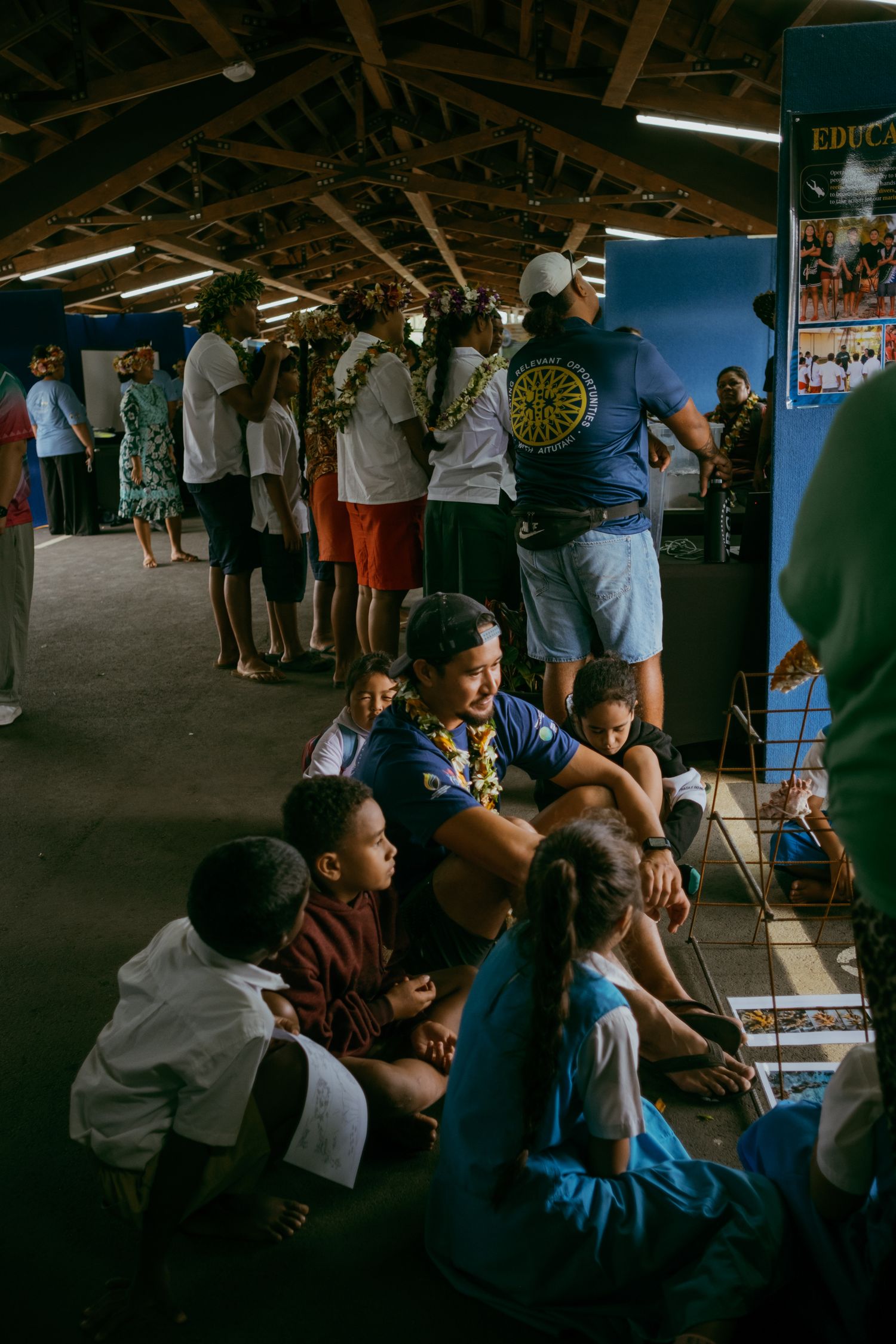 Adults and children gather inside a wooden community hall during a cultural event in a tropical setting.