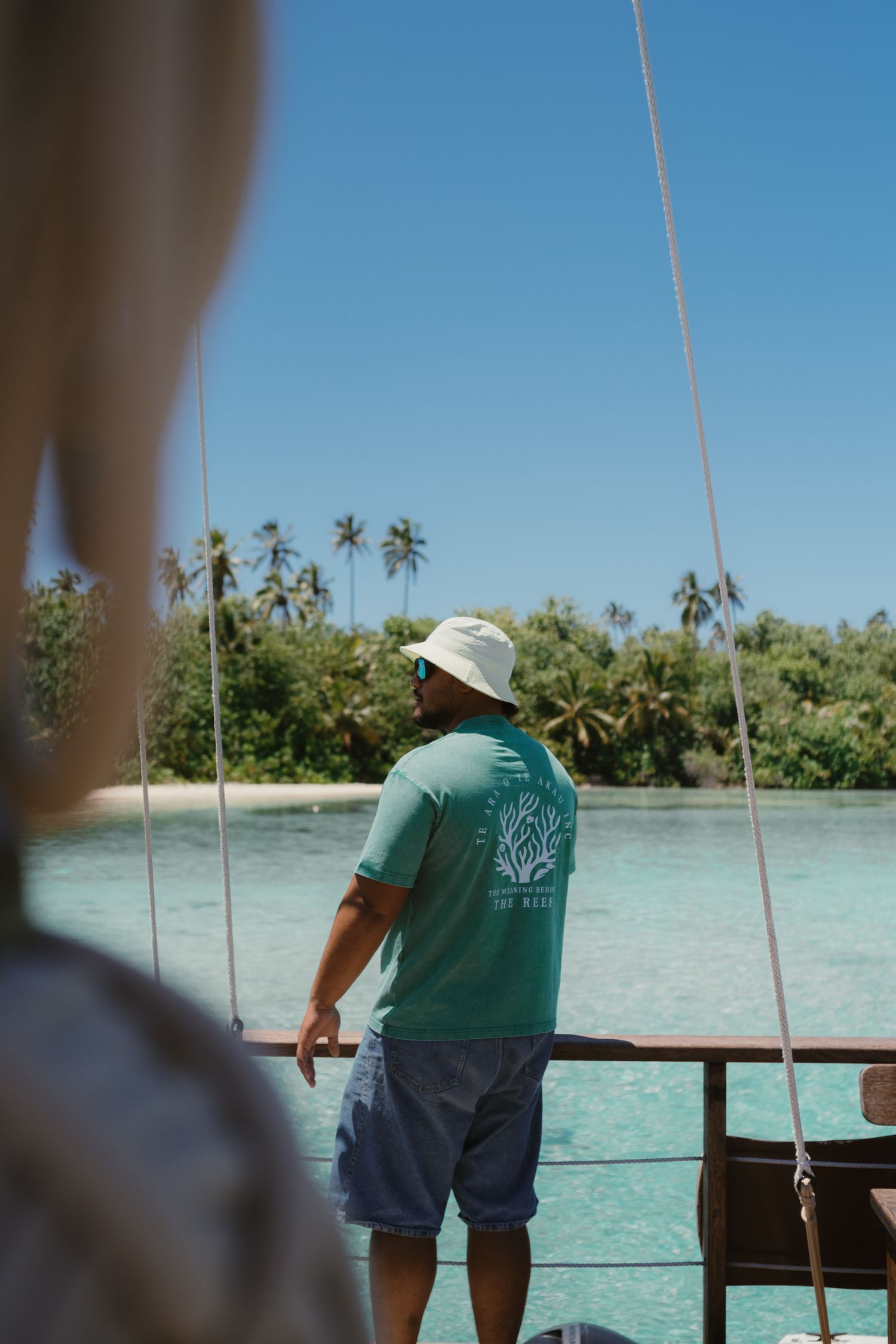 Man in green tee and white hat stands on boat deck gazing at tropical island with palm trees and turquoise water.