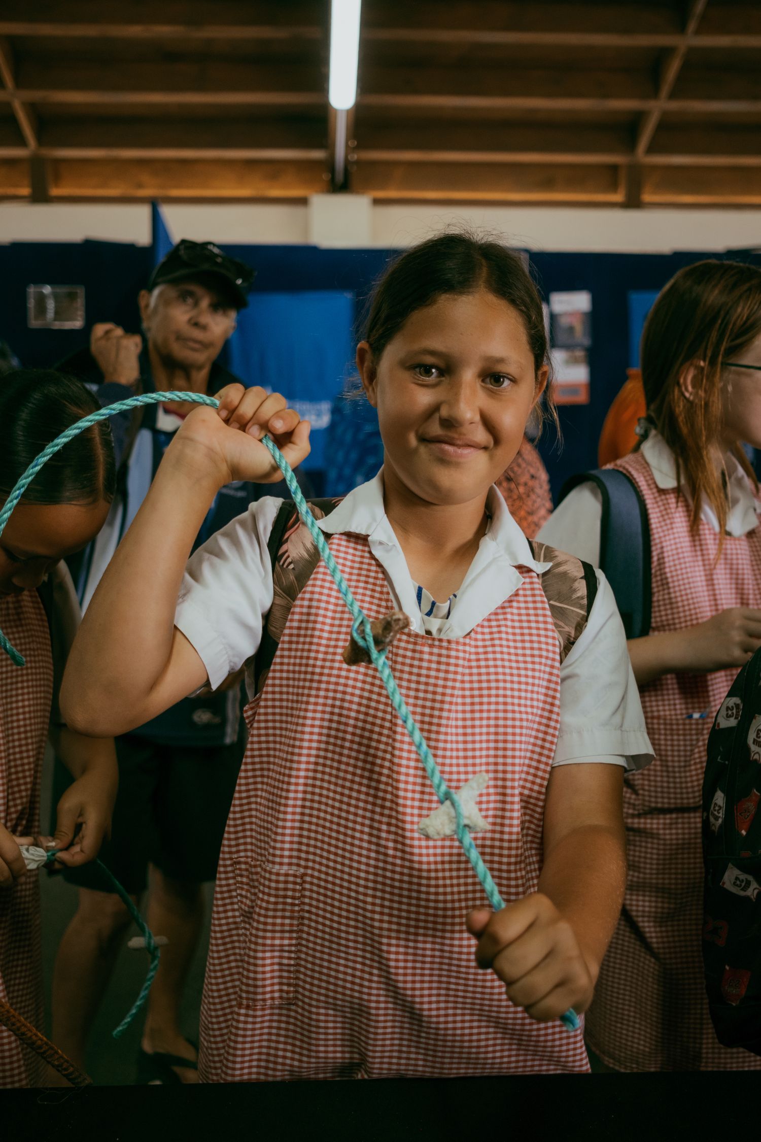 Young schoolgirl in red gingham uniform smiling while holding a turquoise jump rope indoors.