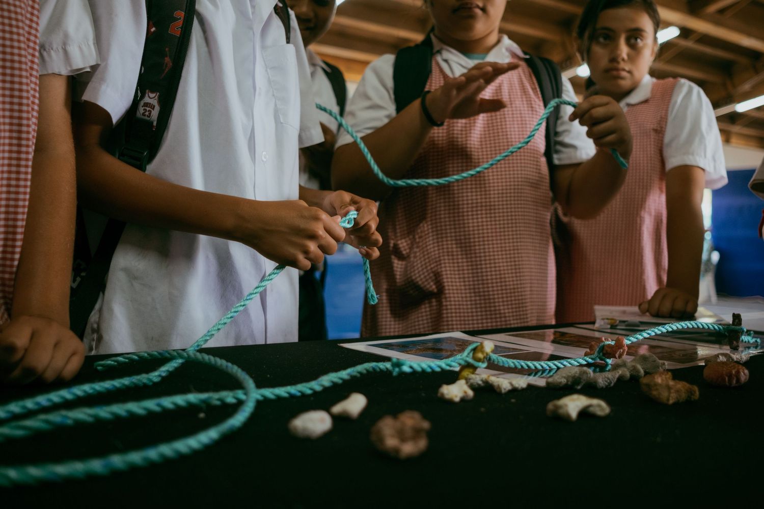 Students in school uniforms handle teal rope and natural objects on a table during an educational hands-on activity.