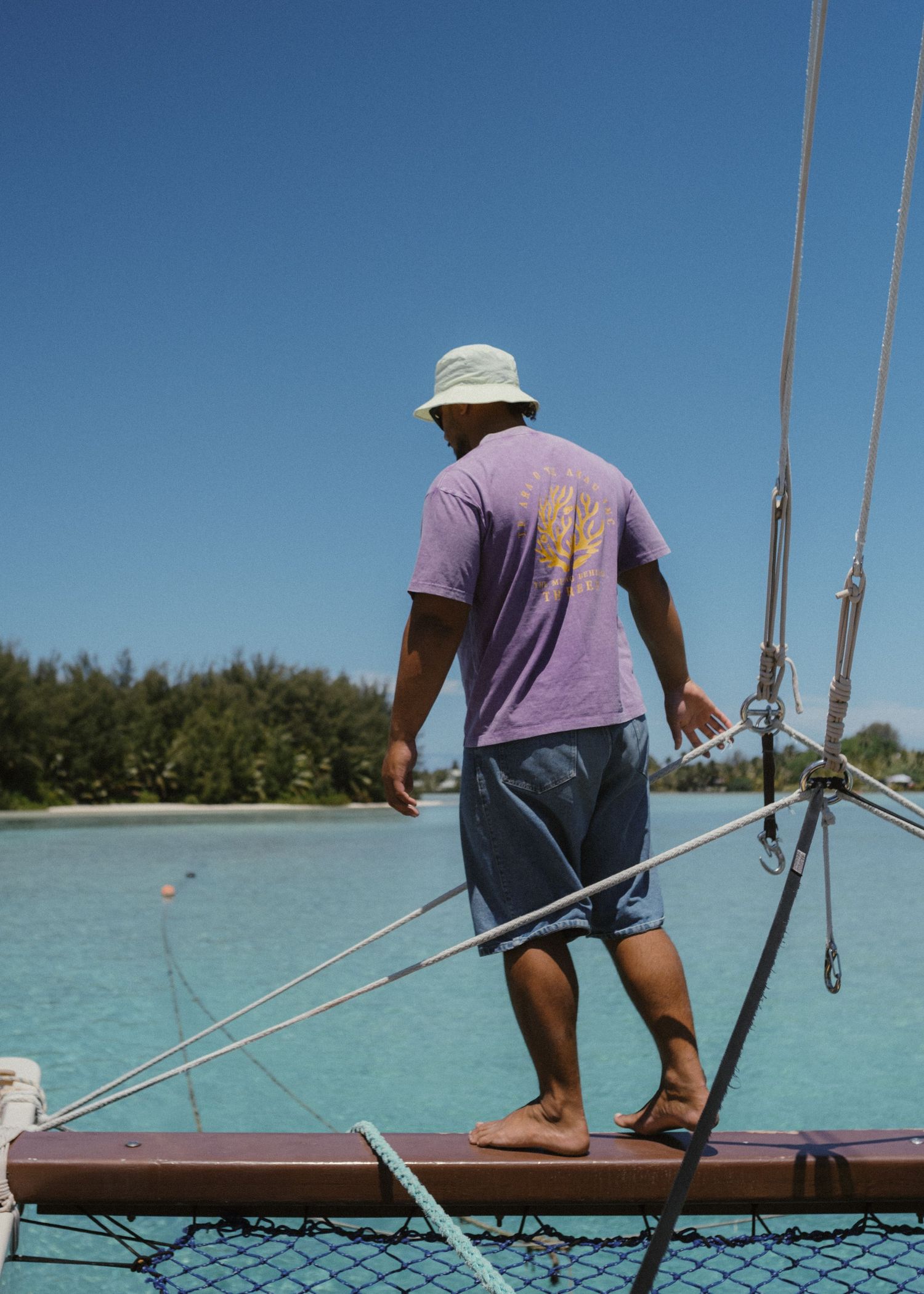 A barefoot man in a purple shirt and bucket hat stands on a sailboat boom over turquoise tropical waters.