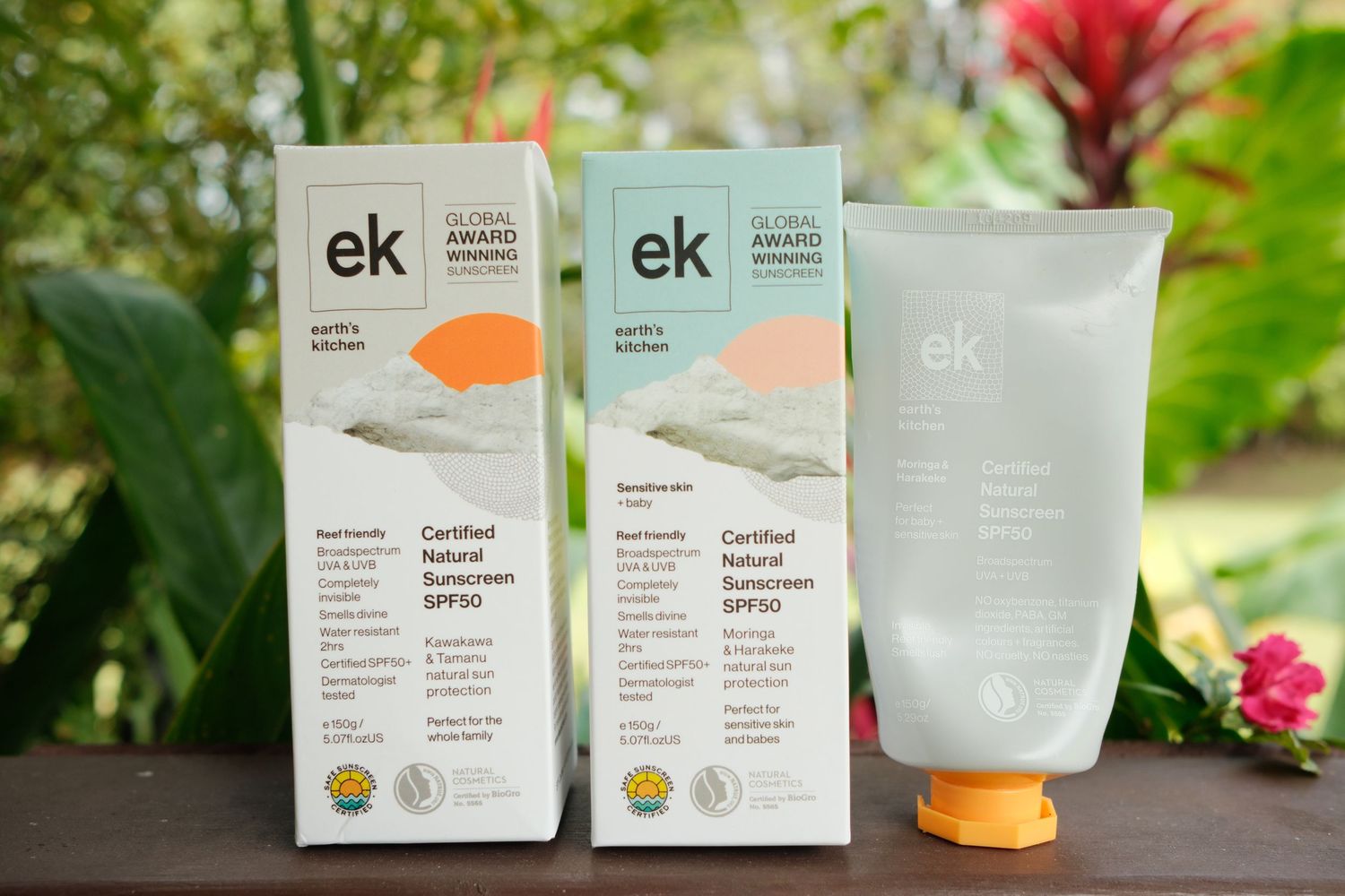 Two EK Certified Natural Sunscreen SPF50 tubes with eco-friendly packaging displayed outdoors among tropical greenery.