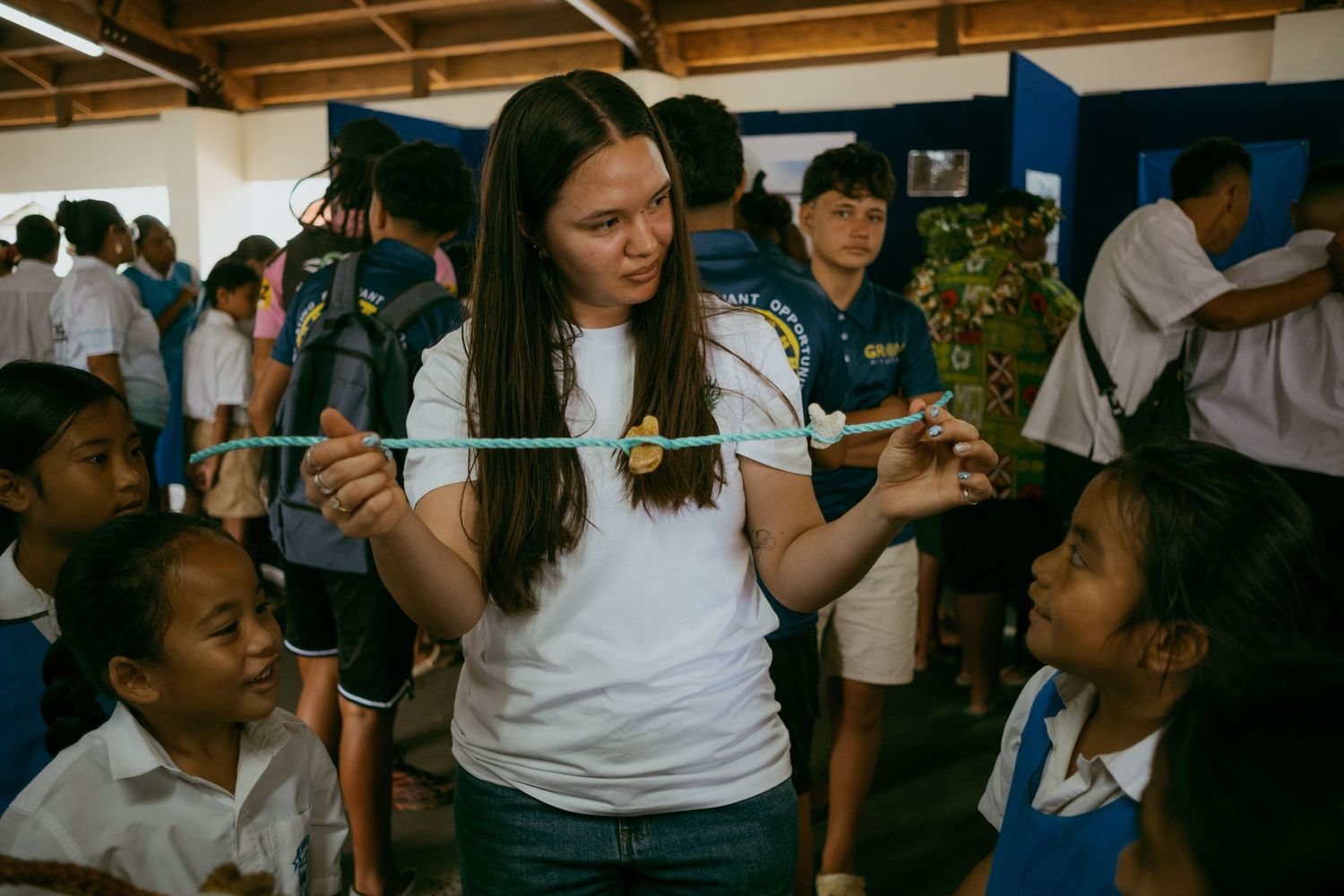 Young woman in white t-shirt engages children in a classroom activity using a measuring tape outdoors.