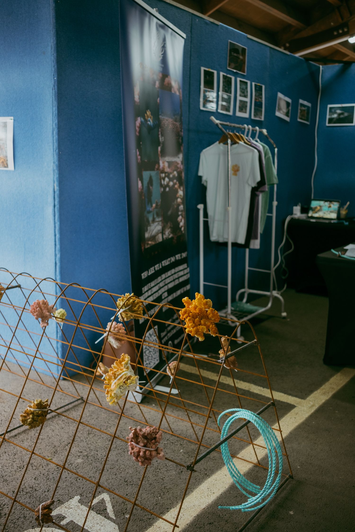 Blue-walled art booth displaying white t-shirts on a rack, decorative wire fence with yarn flowers, and framed photos on the wall.