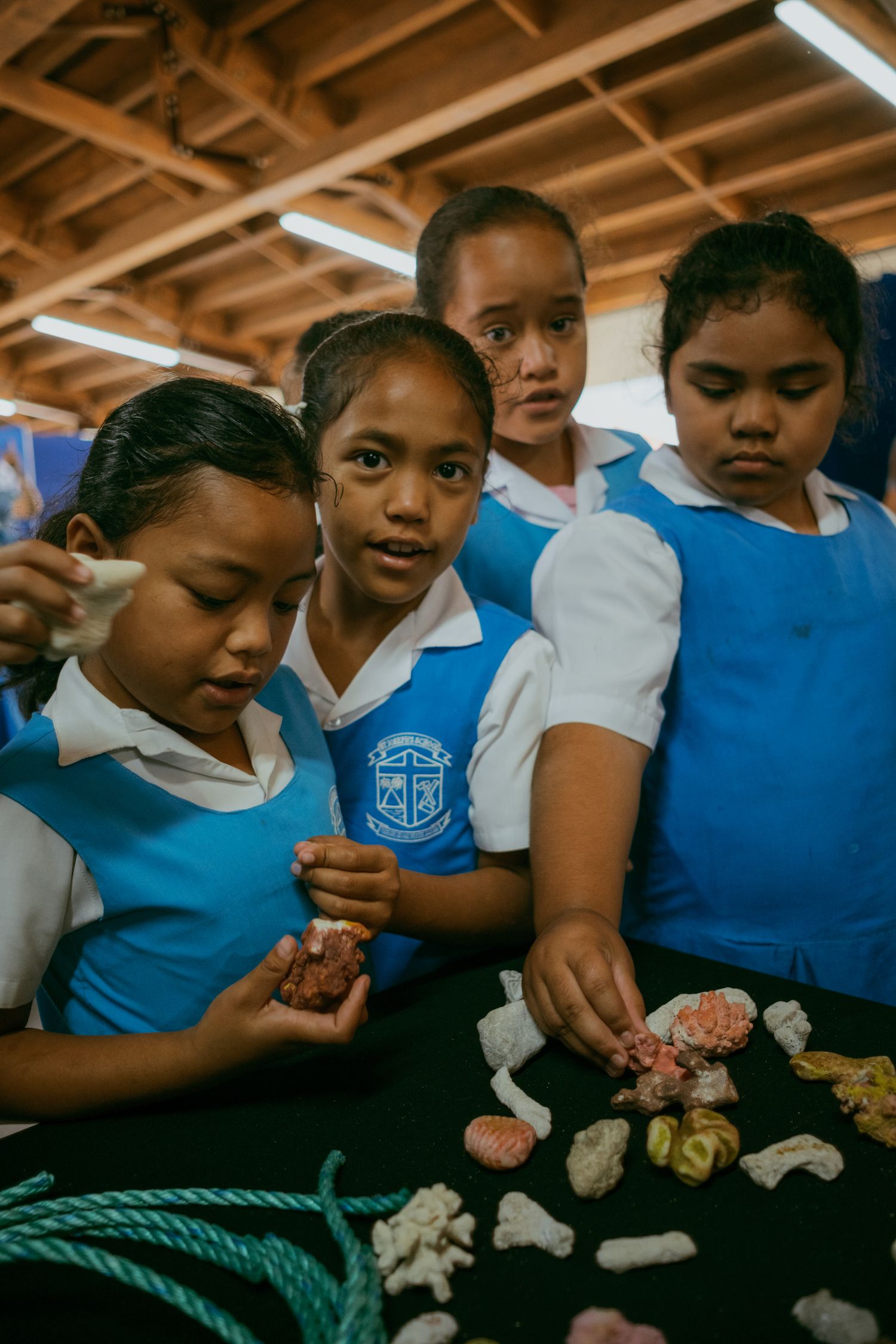 Four schoolgirls in blue uniforms examine coral specimens on a table during an educational marine biology lesson.