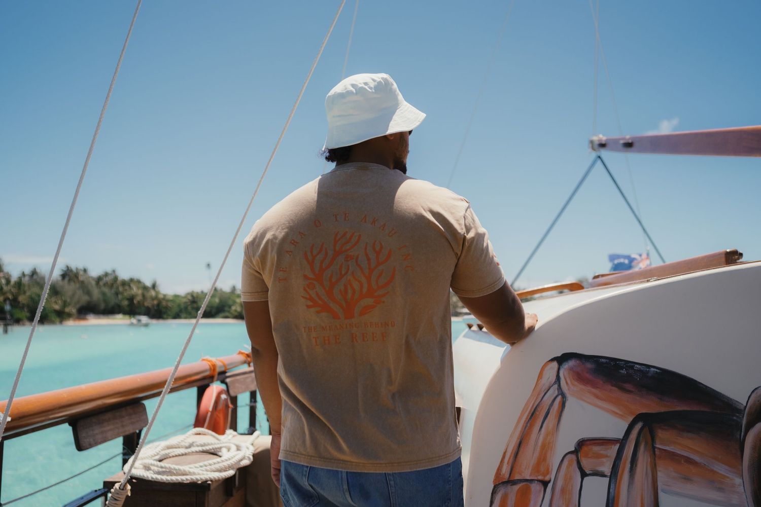 Man in beige graphic tee and white bucket hat standing on a wooden sailboat with turquoise waters.