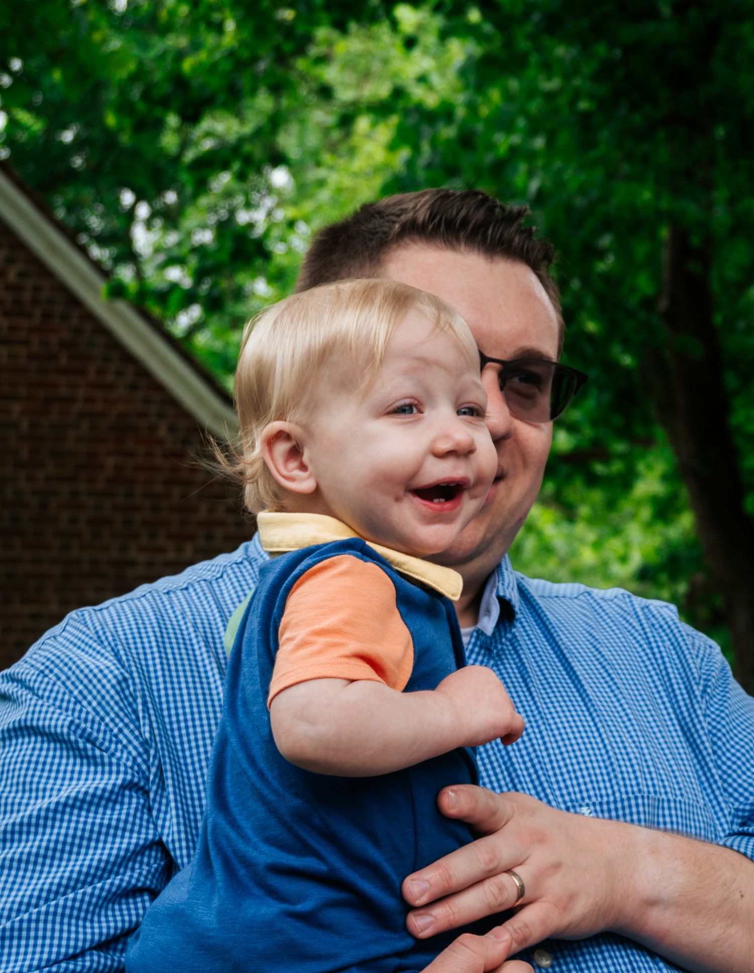 A smiling toddler with blonde hair held by a man in a blue shirt outdoors with green trees in background.