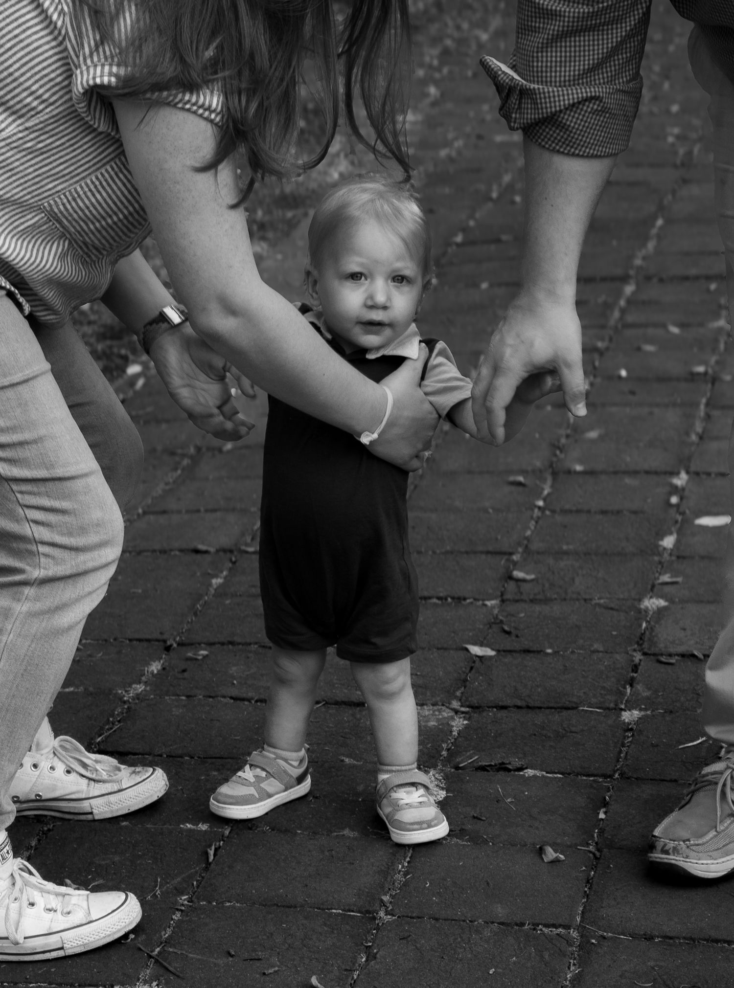 Black and white photo of a toddler learning to walk on brick pavement, supported by two adults holding their hands.