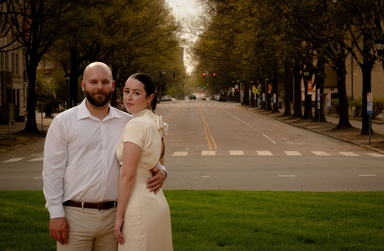 Couple posing on a tree-lined street, man in white shirt and woman in cream dress, urban backdrop.