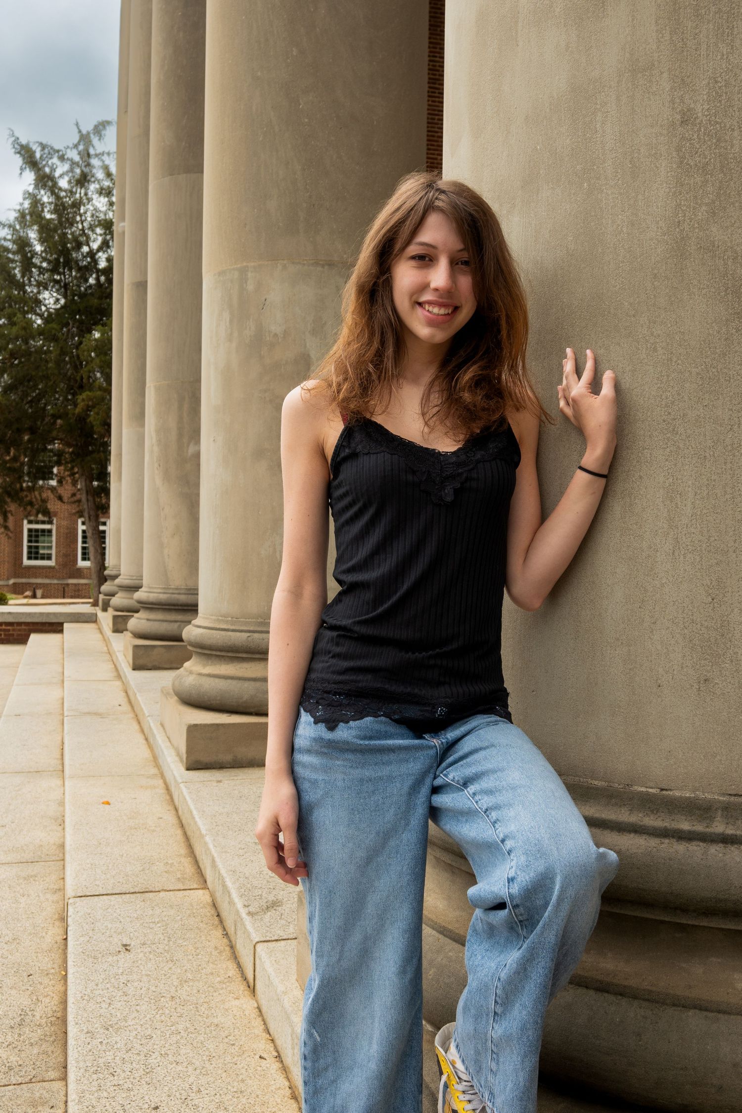 Young woman in black tank top and wide-leg jeans leaning against stone columns outdoors, smiling casually.