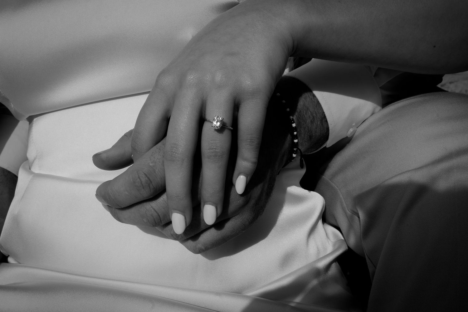 Close-up black and white photo of a couple's hands showing an engagement ring with diamond solitaire.