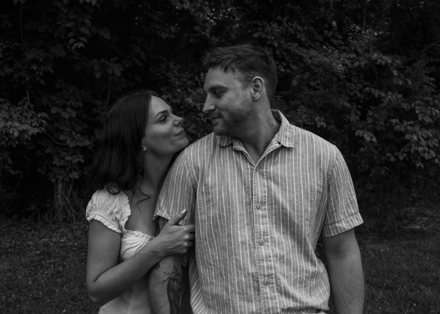 Black and white photo of a smiling couple outdoors, woman leaning on man in striped shirt with trees behind.