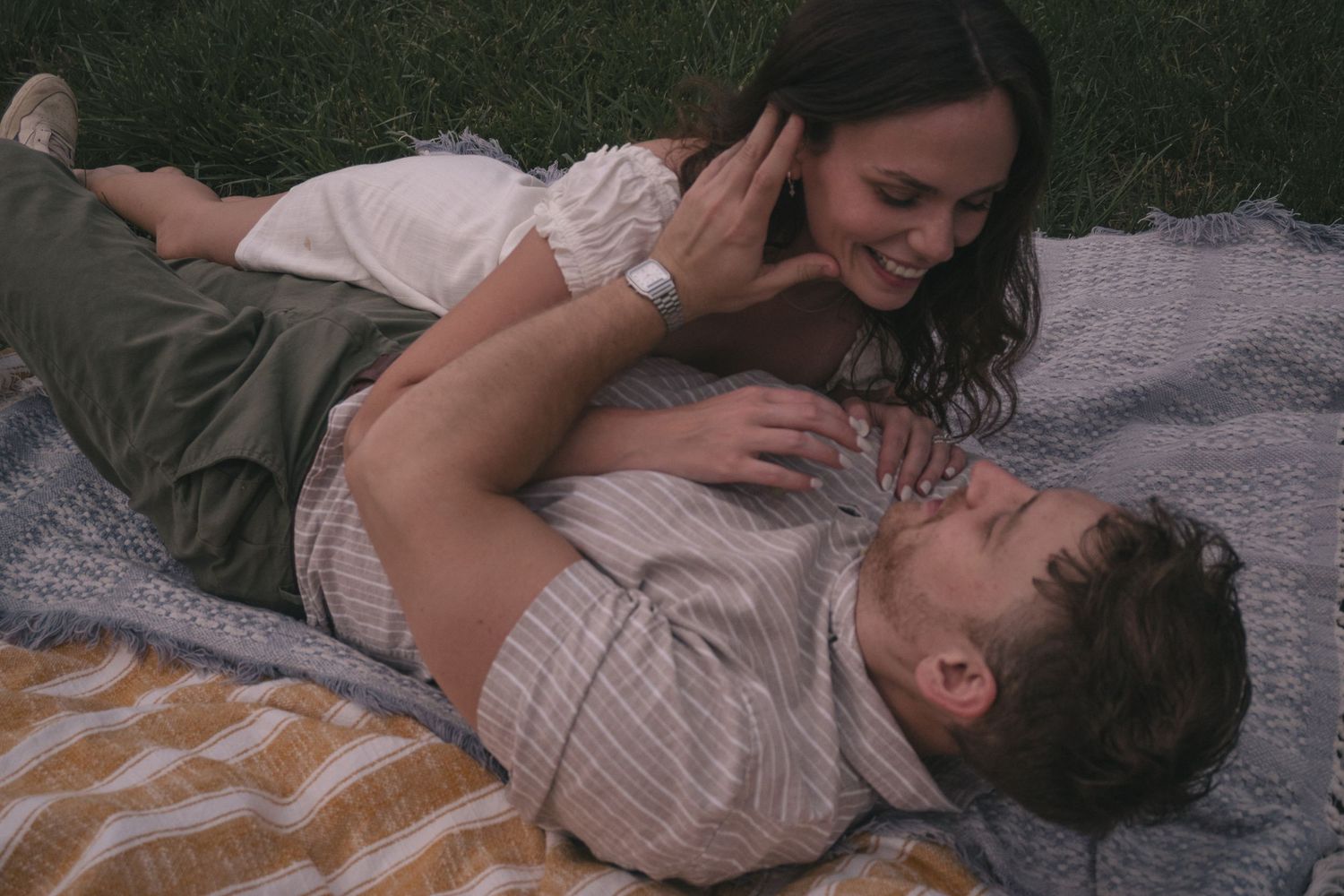 Couple sharing a romantic moment on a blanket in the grass, woman smiling while man lies in her lap.