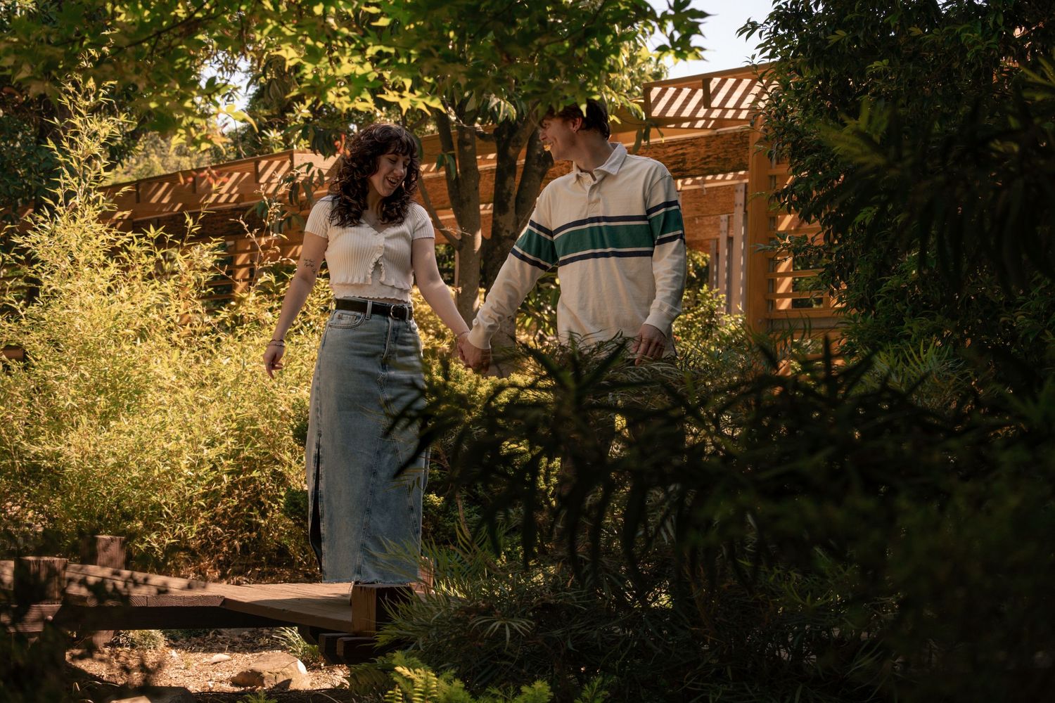 Couple holds hands and smiles at each other on a sunny garden path surrounded by vibrant green foliage.