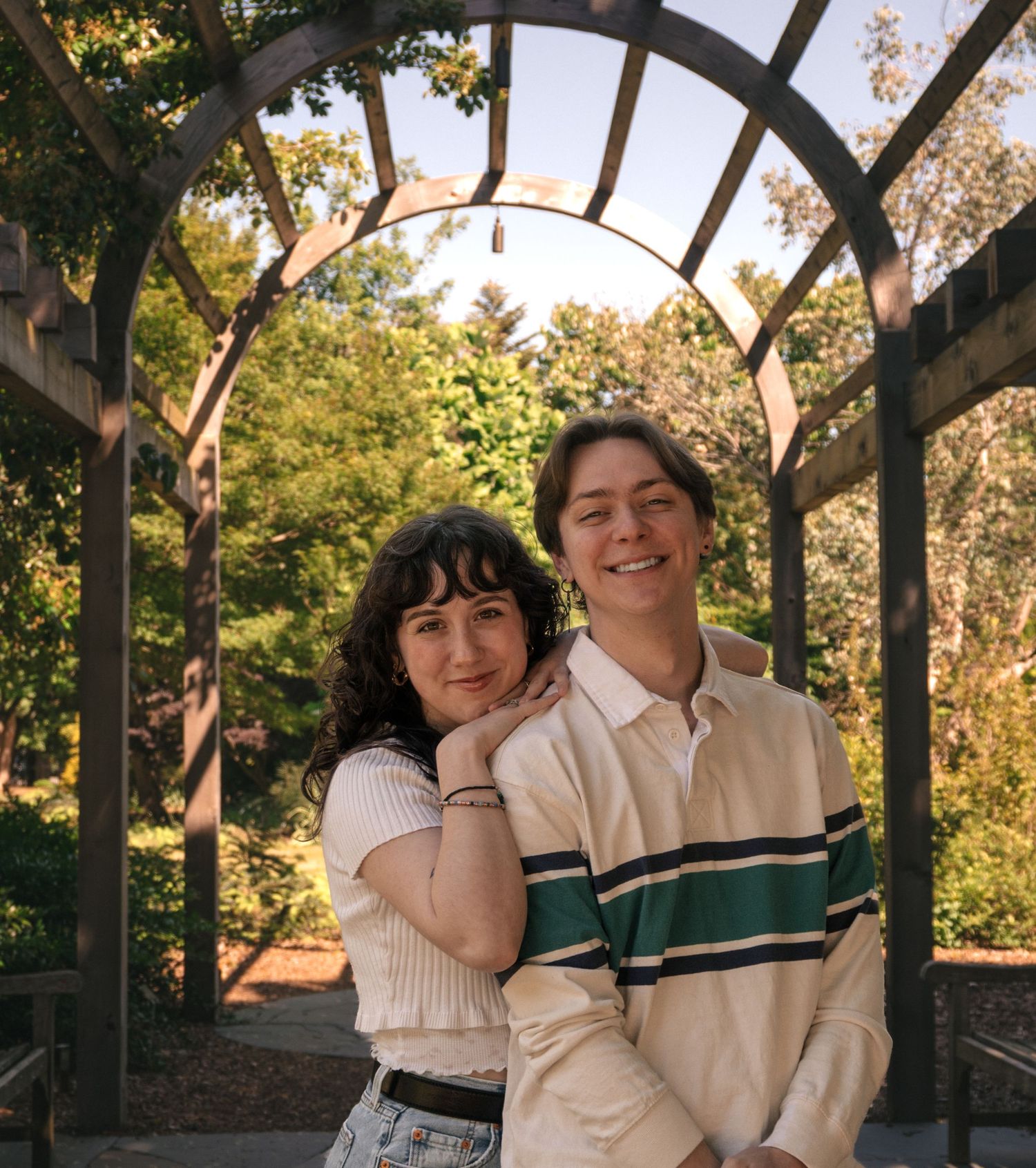 A smiling couple poses under a wooden garden arch surrounded by lush green trees on a sunny day.