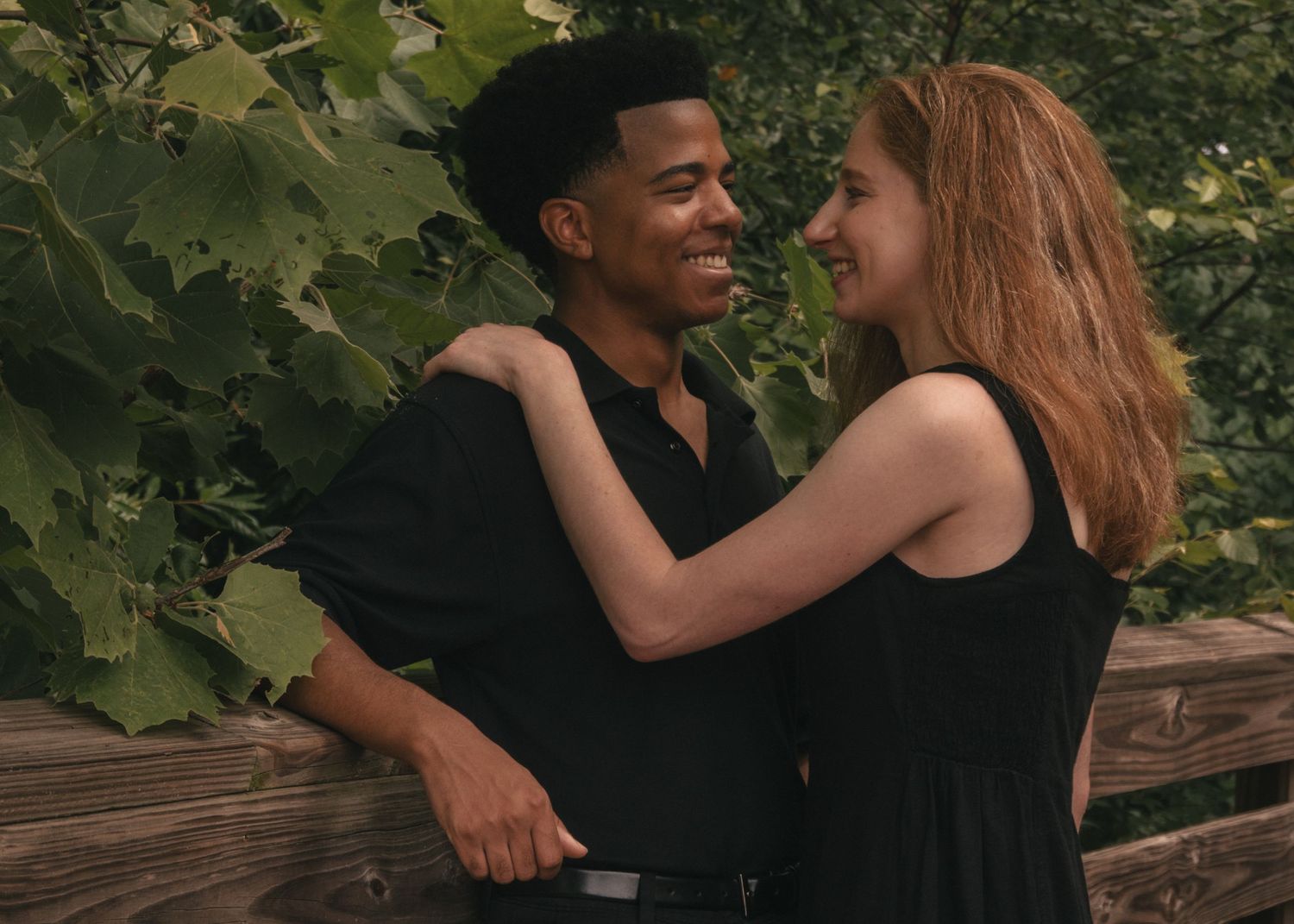 A smiling interracial couple in black outfits embracing outdoors near a wooden fence with lush green foliage.