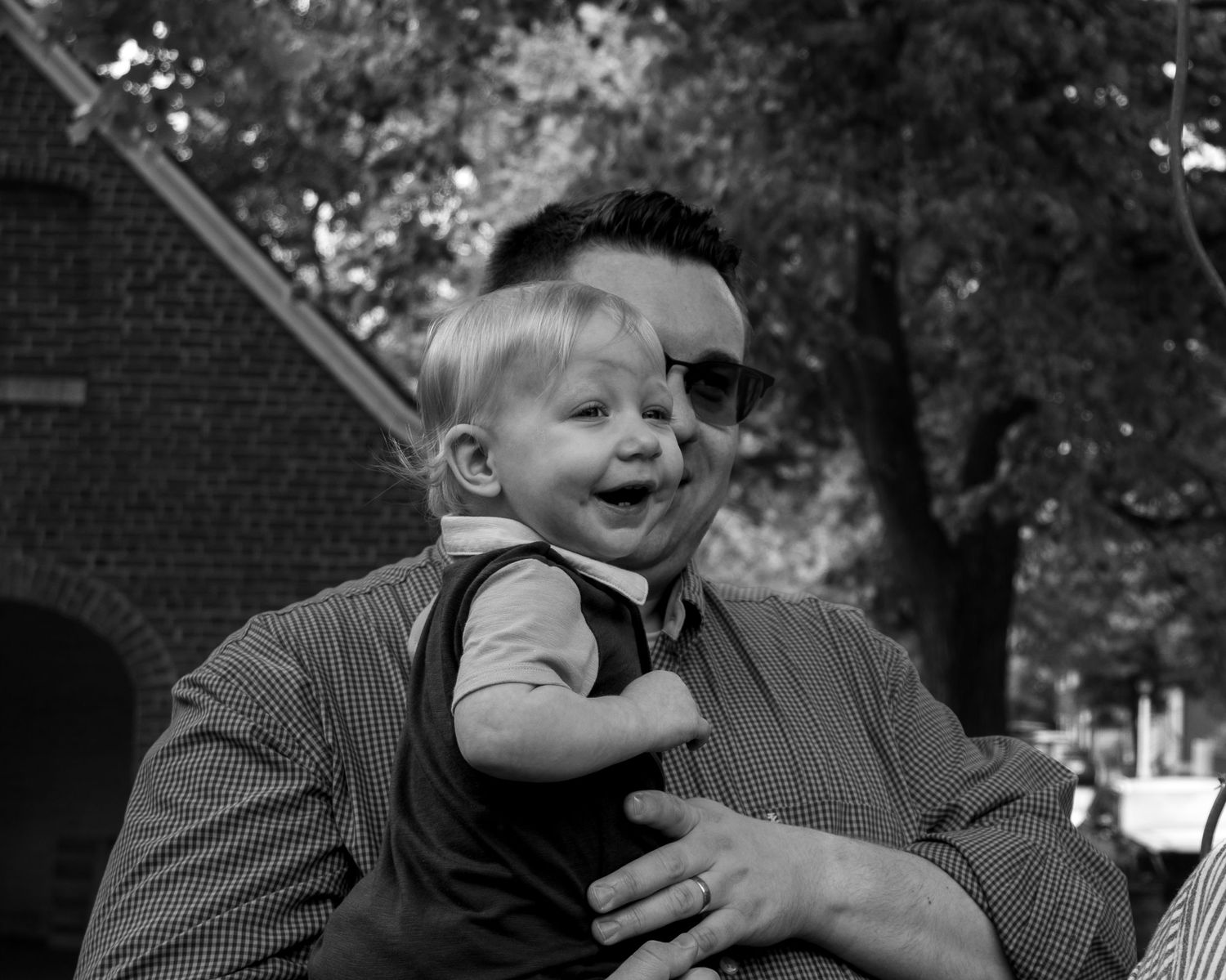 Black and white portrait of a smiling baby being held by an adult outdoors.