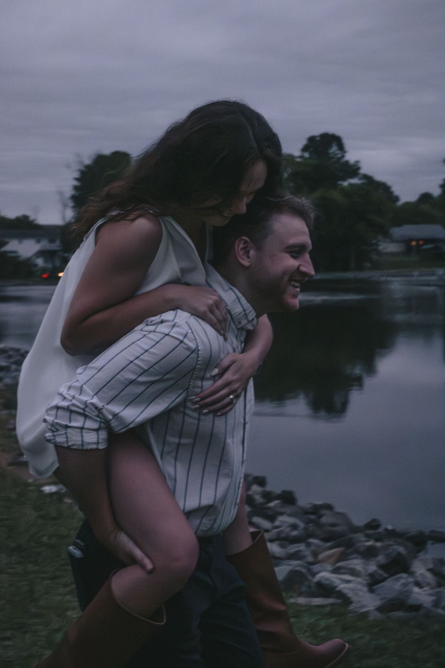 Joyful man giving a laughing woman a piggyback ride near a reflective lake at dusk.
